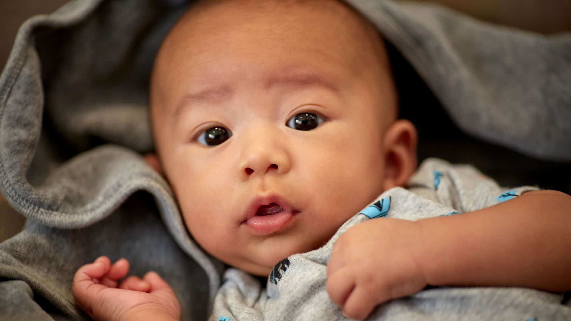 A close-up of a baby lying down, wrapped in a soft grey hoodie, with wide, expressive eyes and a slightly open mouth in a candid moment of curiosity.