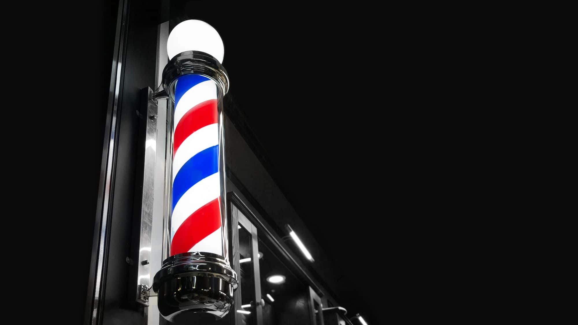 A brightly lit traditional barbershop pole with red, white, and blue spirals glowing against a sleek black night backdrop.