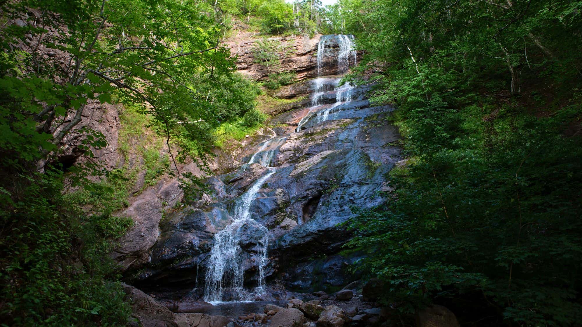 A tall, cascading waterfall descends in thin streams down a rugged, dark cliff face surrounded by dense green foliage, creating a dramatic scene in the forested landscape.
