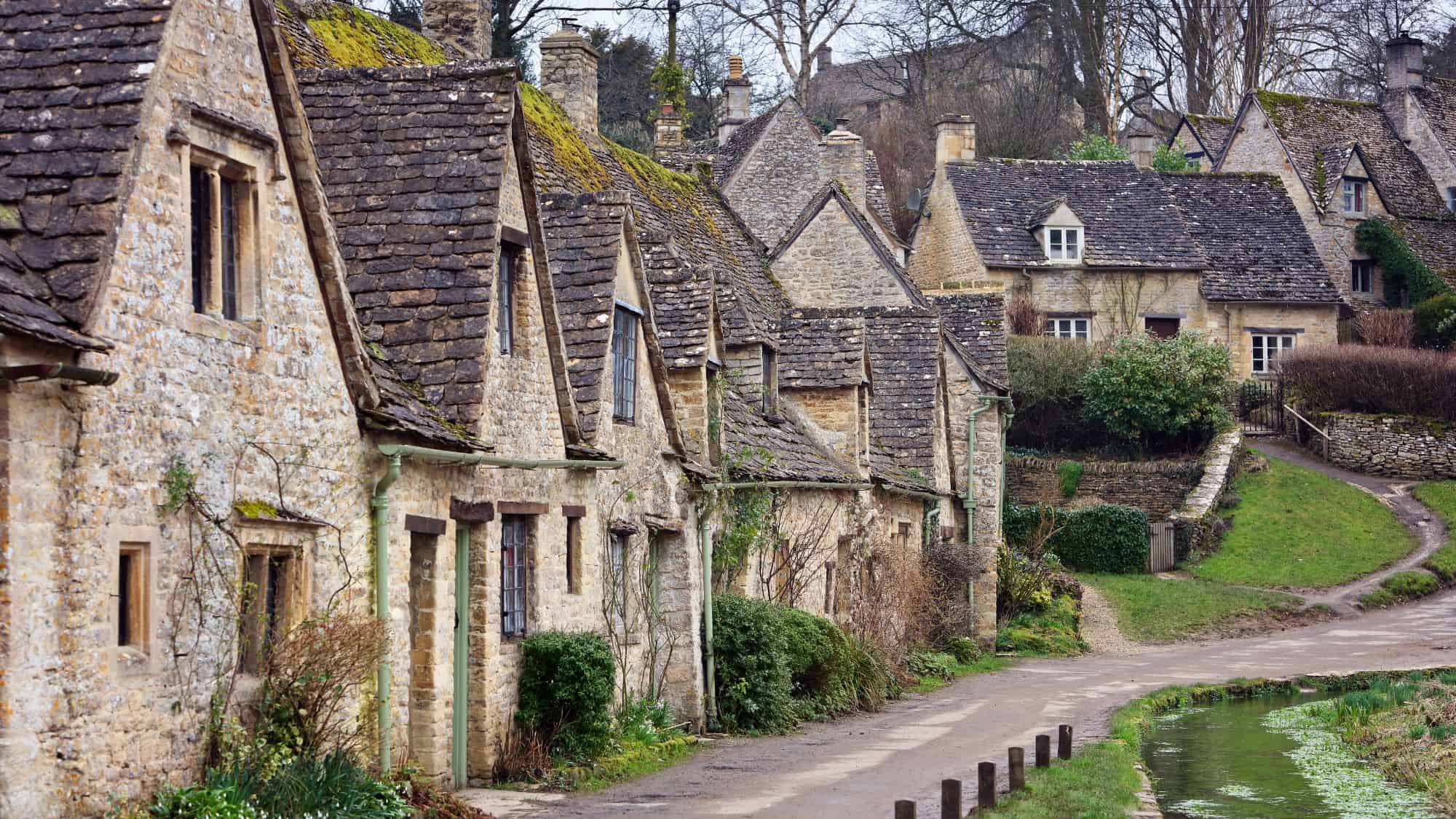 A row of charming stone cottages with steep slate roofs, covered in moss and surrounded by lush gardens, lines a narrow village path beside a tranquil stream.