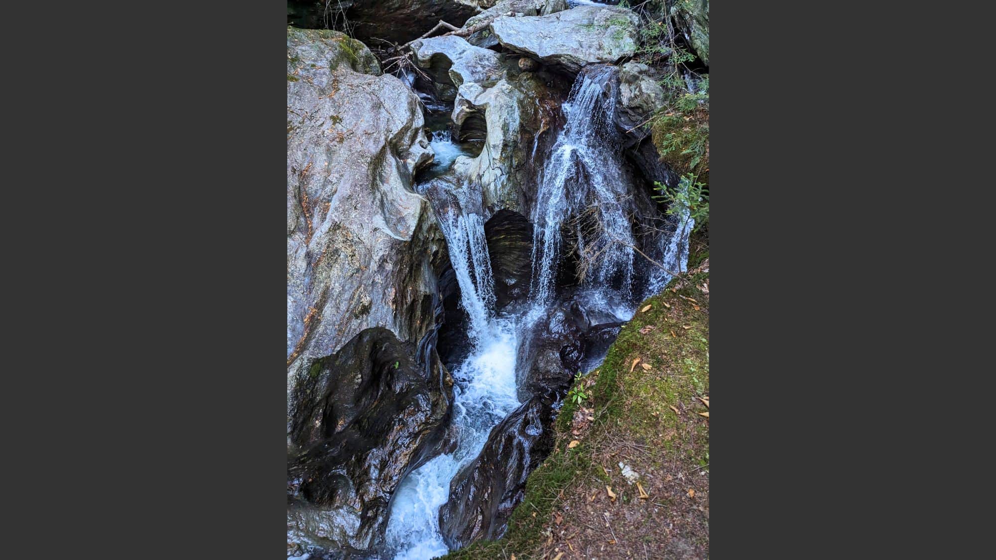 A compact and powerful waterfall pours into a chiseled, narrow rocky gorge, framed by sculpted stone walls and surrounded by forest undergrowth, showcasing nature’s raw and intricate carving work.