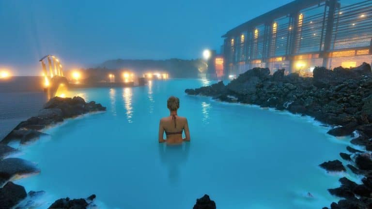 A woman wades in the milky blue geothermal waters of Iceland’s Blue Lagoon at night, surrounded by mist, dark volcanic rocks, and softly glowing lamps that reflect off the water.