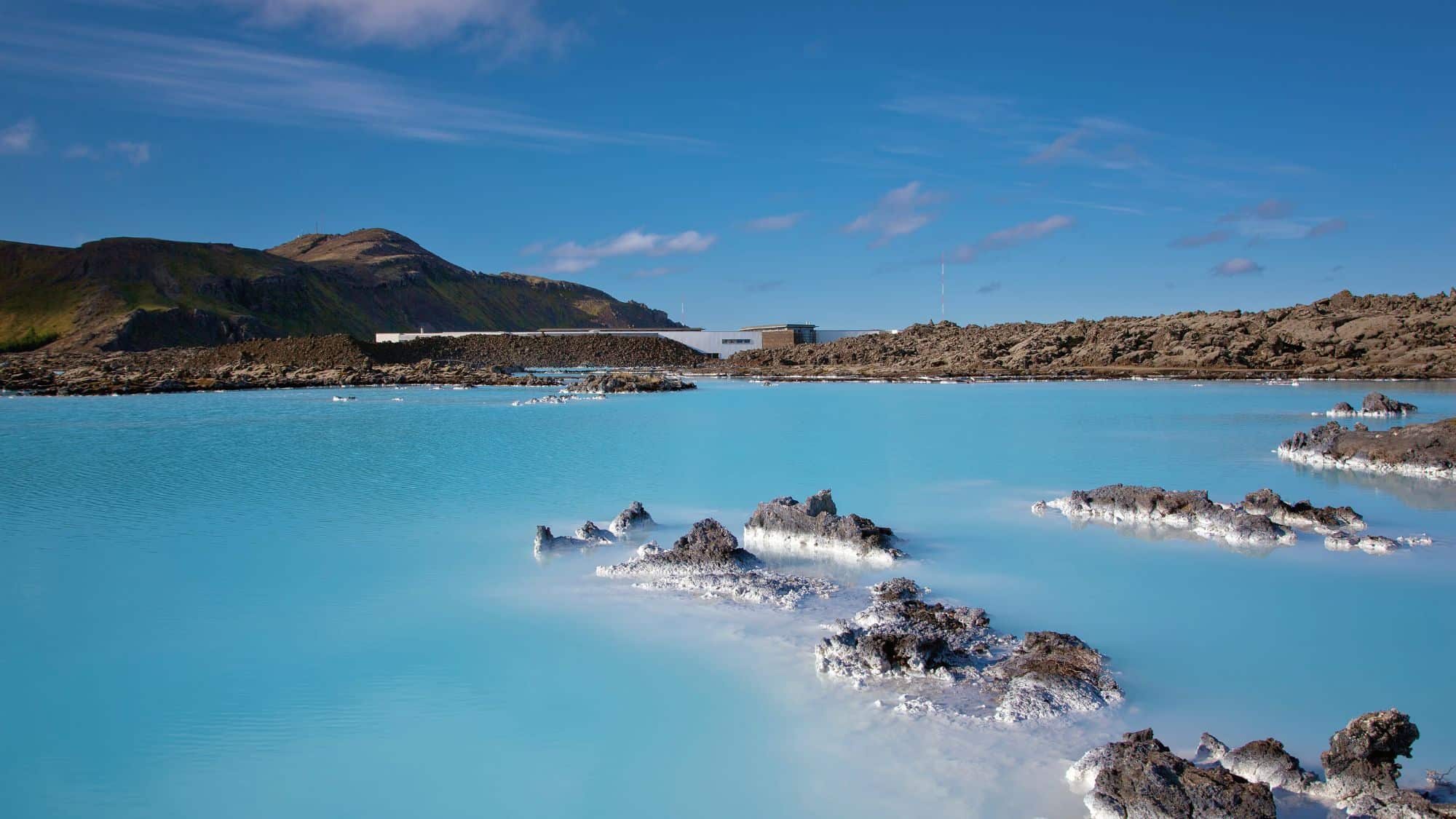 Bright blue mineral-rich waters of Iceland’s Blue Lagoon contrast against dark volcanic rock formations under a clear sky, with distant spa buildings nestled into the landscape.
