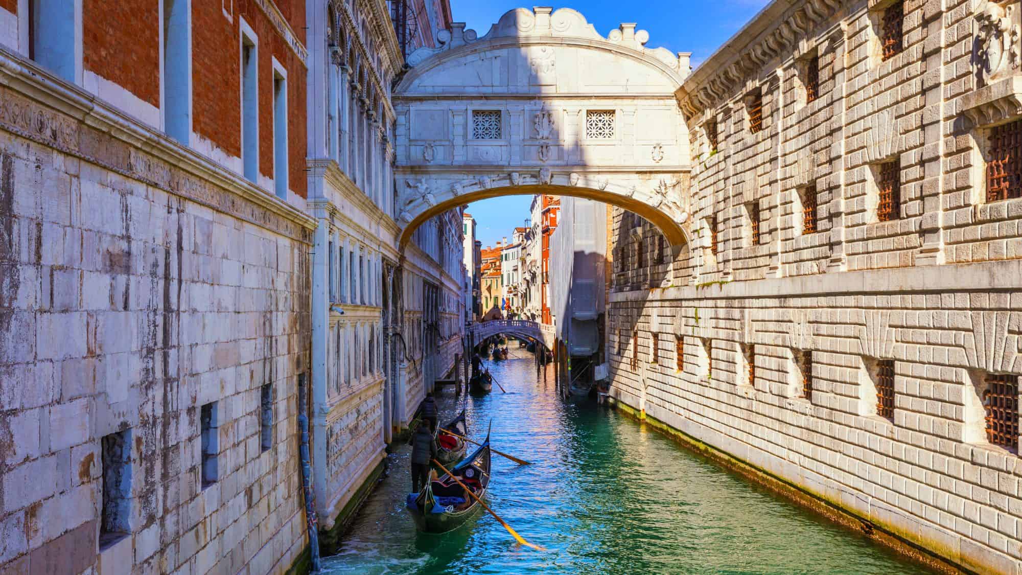 The white limestone Bridge of Sighs spans a narrow canal in Venice, with gondolas gliding below between historic stone buildings.