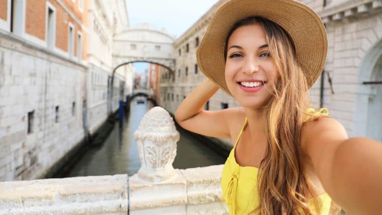 A woman in a yellow dress and straw hat takes a cheerful selfie on a bridge in Venice, with the iconic white Bridge of Sighs and a canal in the background.