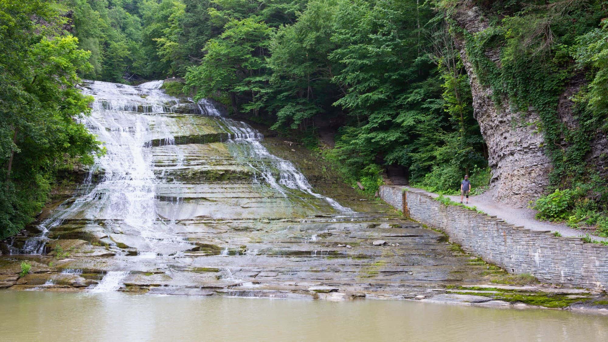 A broad, tiered waterfall flows gently down a long rock face with a pedestrian trail running alongside, flanked by dense greenery and a towering rock wall.