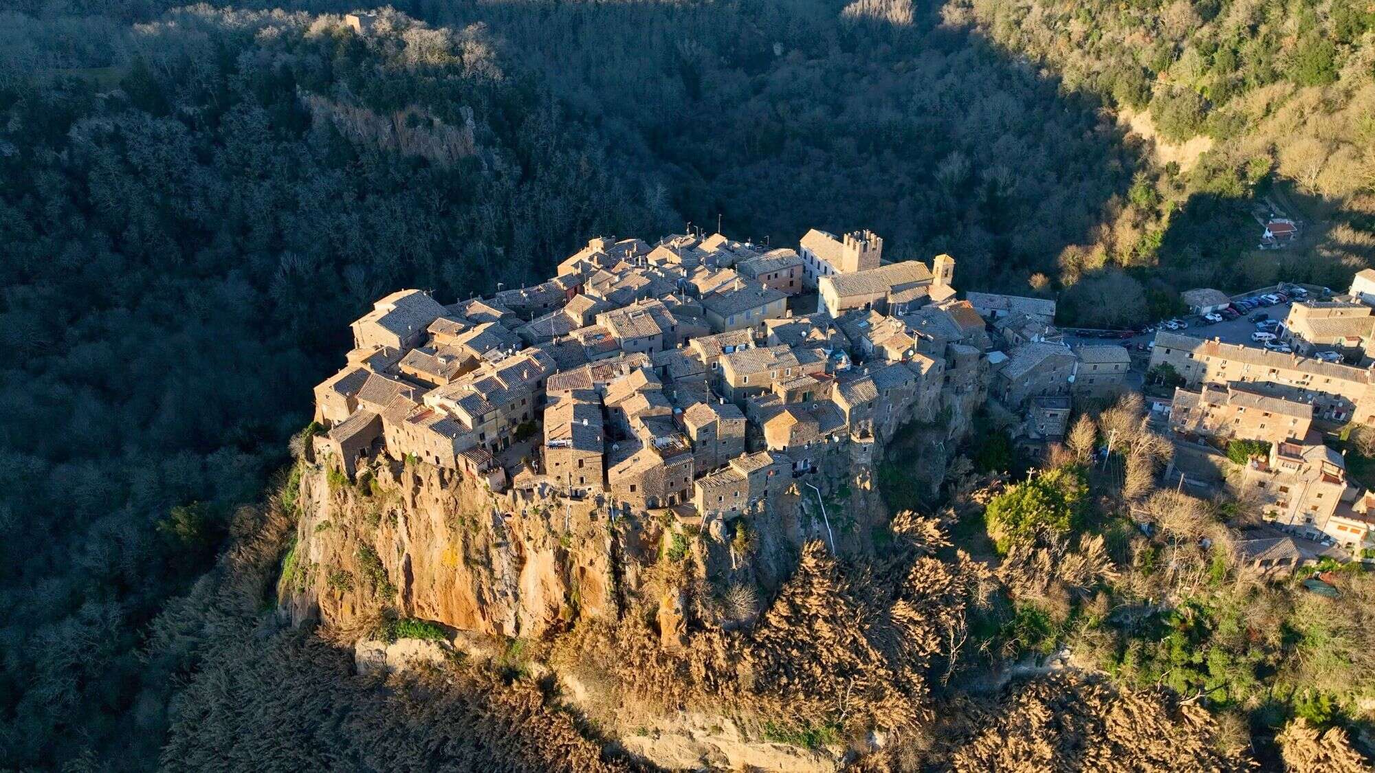 A compact cluster of ancient stone houses perched dramatically on a cliffside, surrounded by dense forest, bathed in warm golden light.