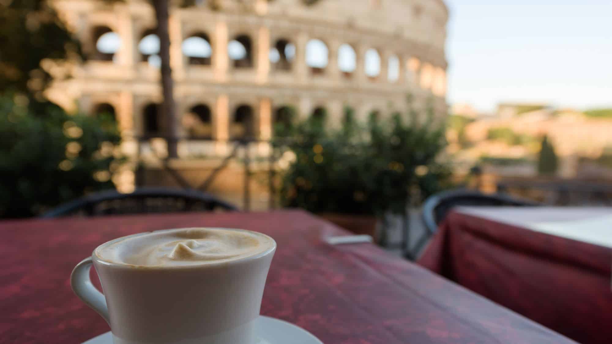 A foamy cappuccino sits on a red café table in the foreground, with the blurred arches of Rome’s Colosseum rising in the background, evoking a classic Italian morning scene.