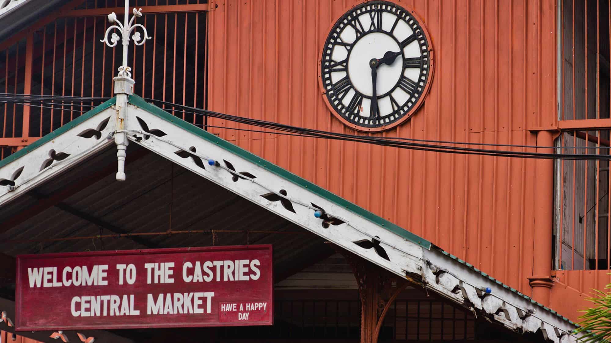 Close-up of the Castries Central Market entrance in St. Lucia, with a red and white sign welcoming visitors and an old-fashioned clock mounted above on a corrugated red building.