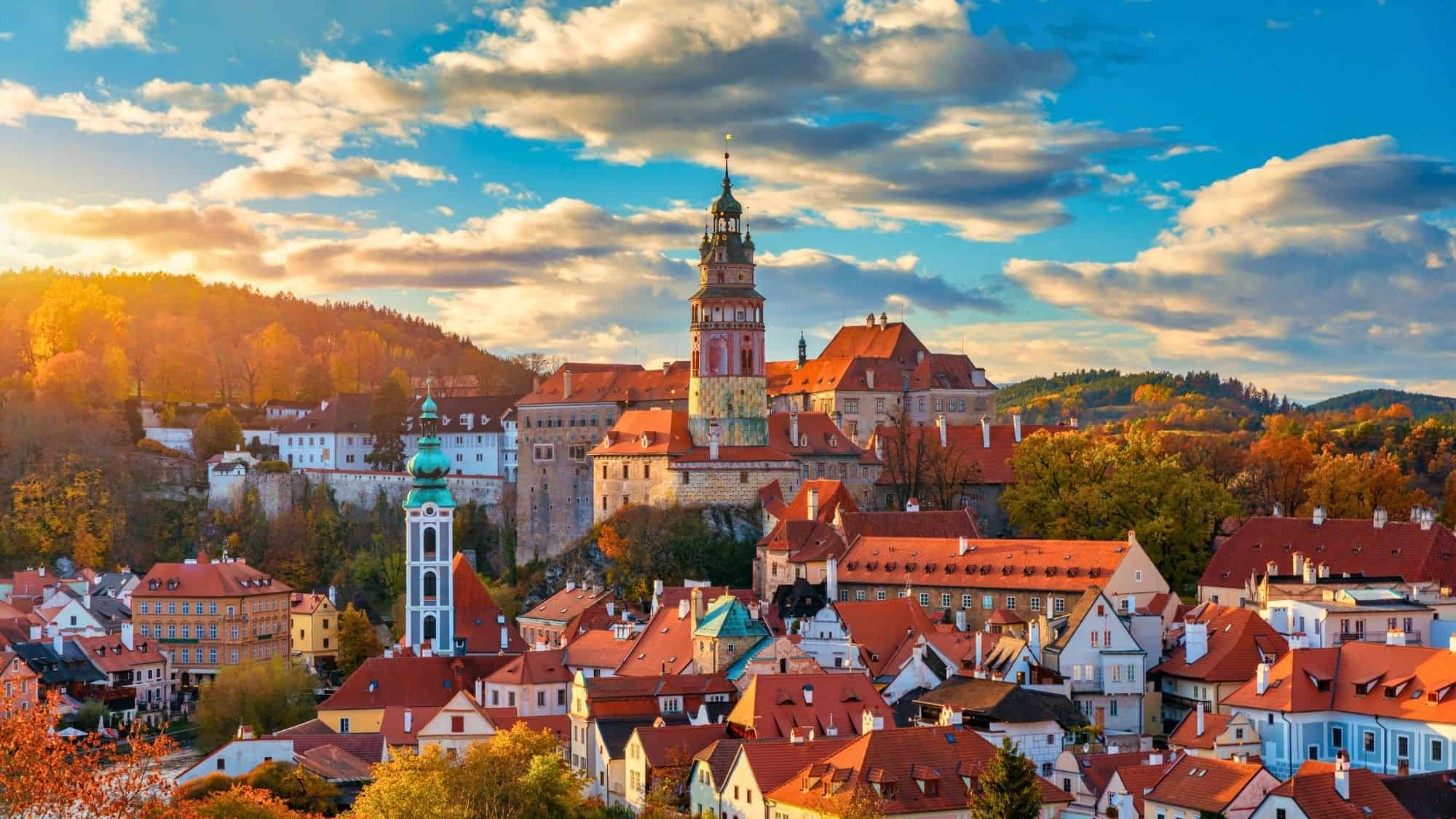 An enchanting medieval town with red-tiled rooftops and a striking castle tower, set against a backdrop of autumn-colored trees and dramatic skies.