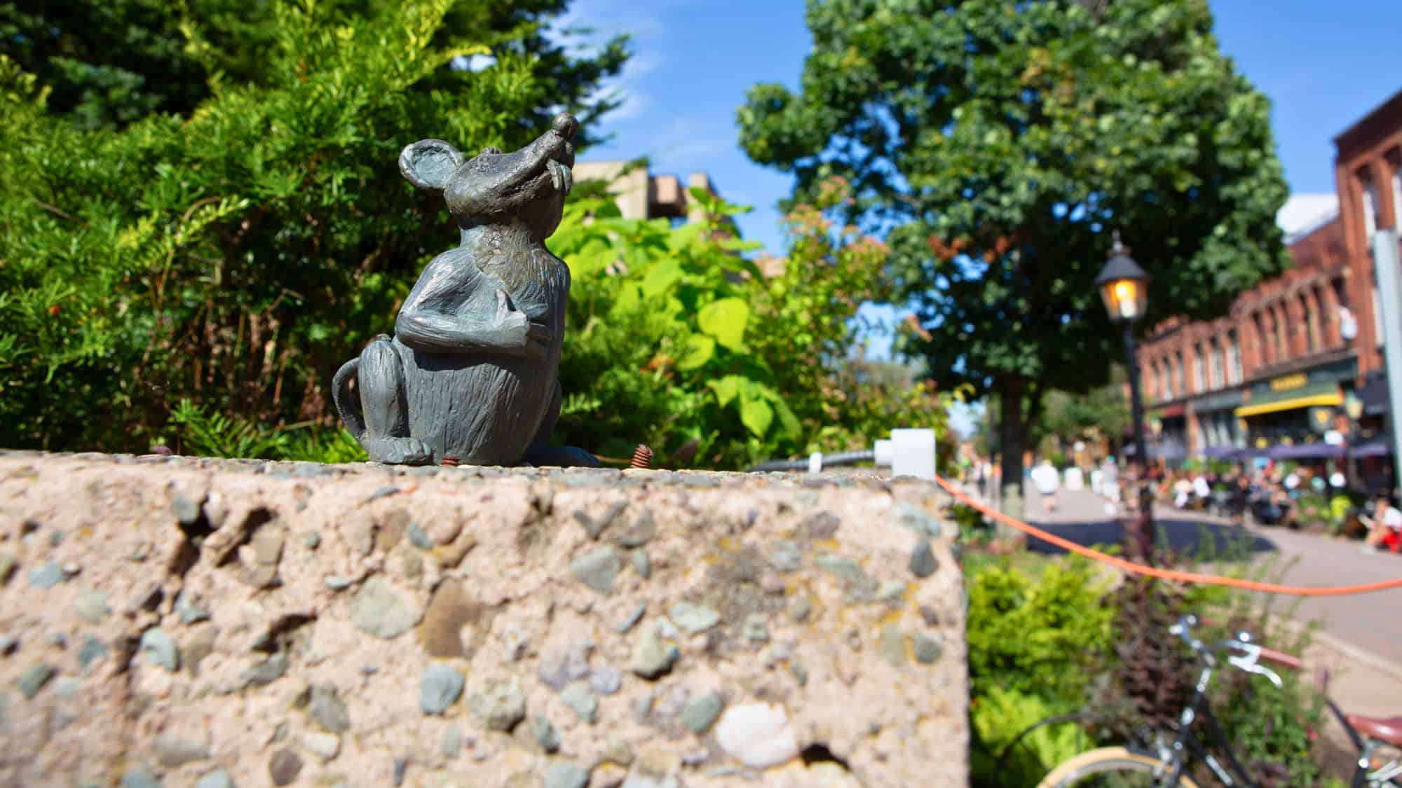 A tiny bronze mouse sculpture sits atop a concrete wall in a lush garden setting, with a vibrant pedestrian street in Charlottetown visible in the blurred background.