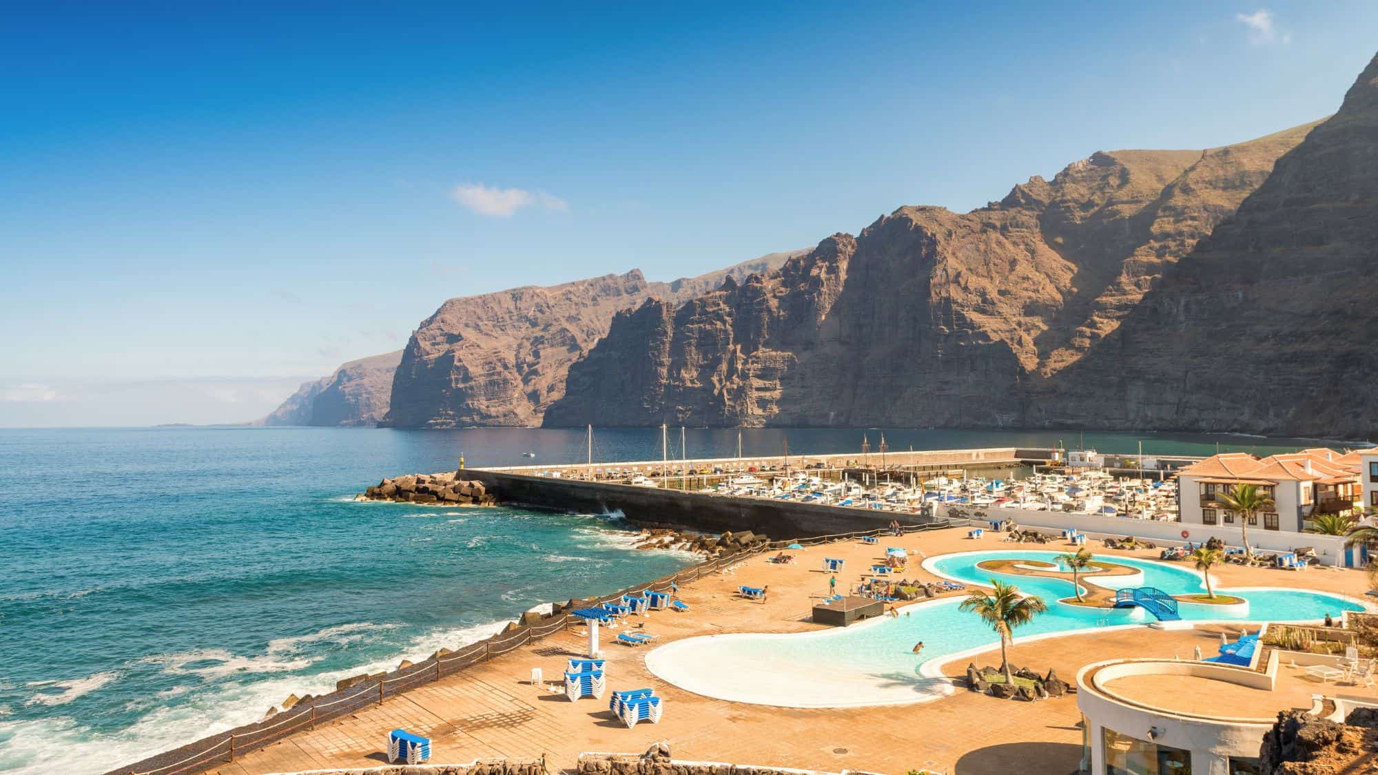 A scenic view of the rugged Los Gigantes cliffs in Tenerife rising sharply from the Atlantic Ocean, with a waterfront resort and curving pool in the foreground beside a marina lined with white boats.