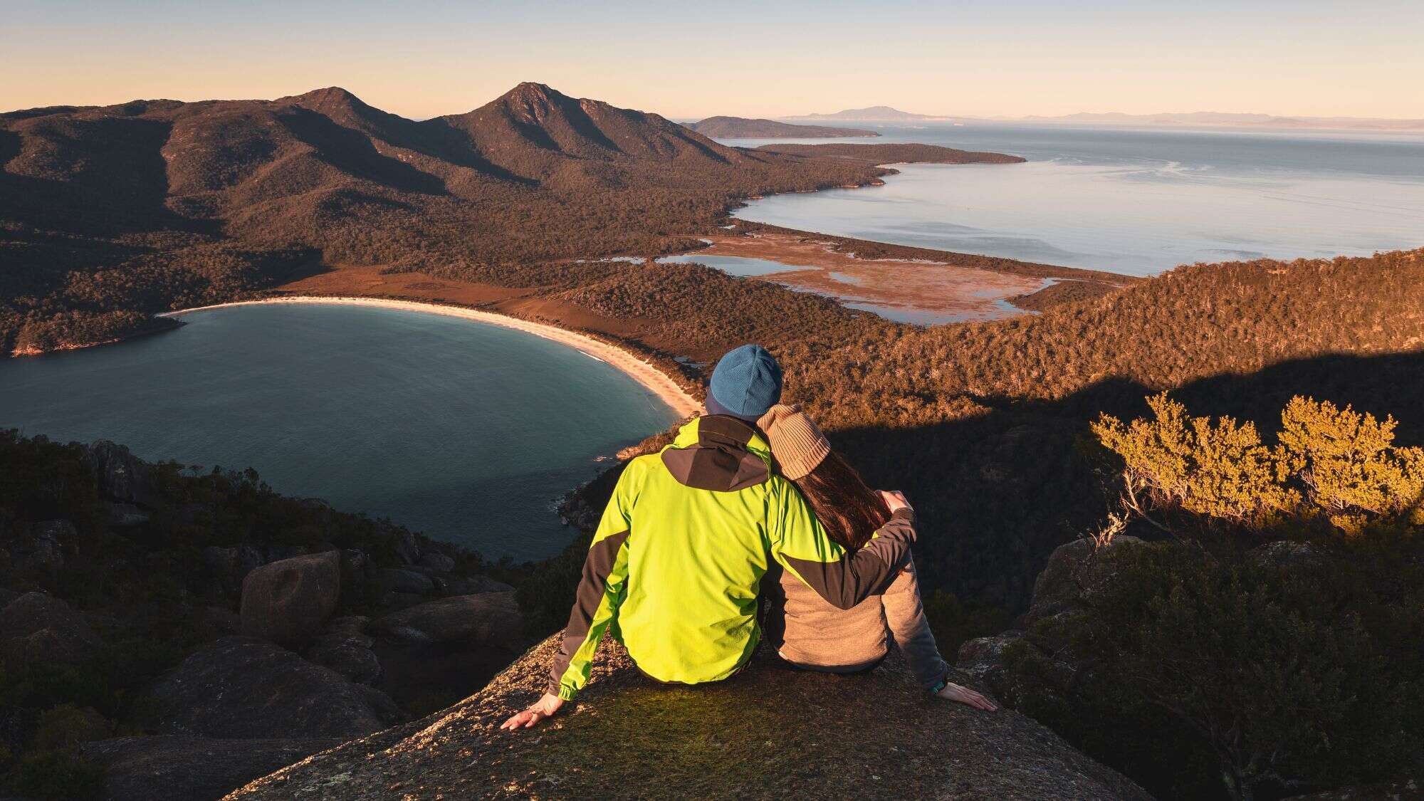 A couple sits embraced on a rocky outcrop, gazing out at the crescent-shaped Wineglass Bay and rugged forested mountains bathed in golden sunlight. The expansive coastline and calm sea suggest peace and awe.