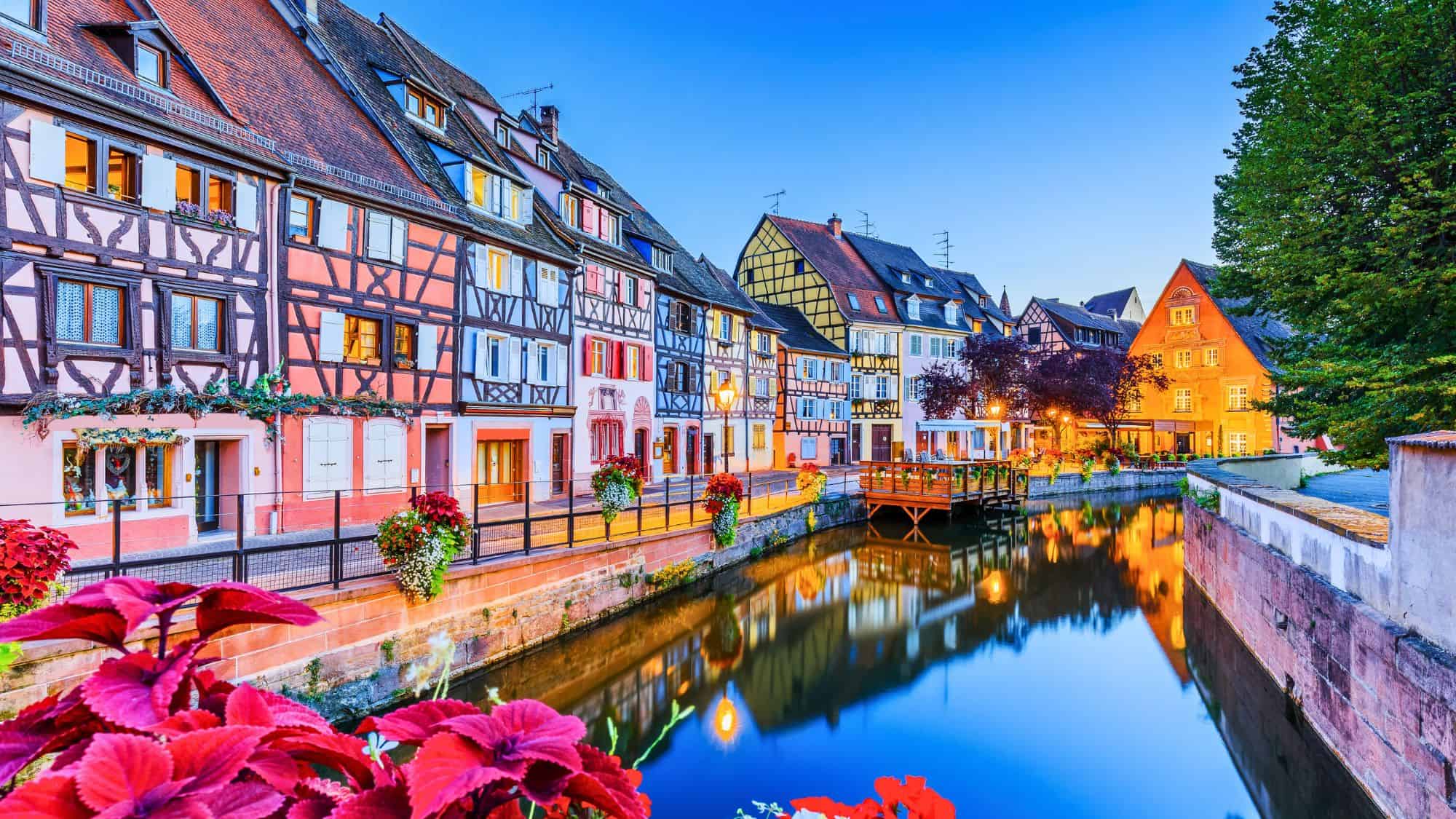 Colorful half-timbered houses reflect in a calm canal at twilight, with flower boxes and streetlights creating a fairy-tale atmosphere.