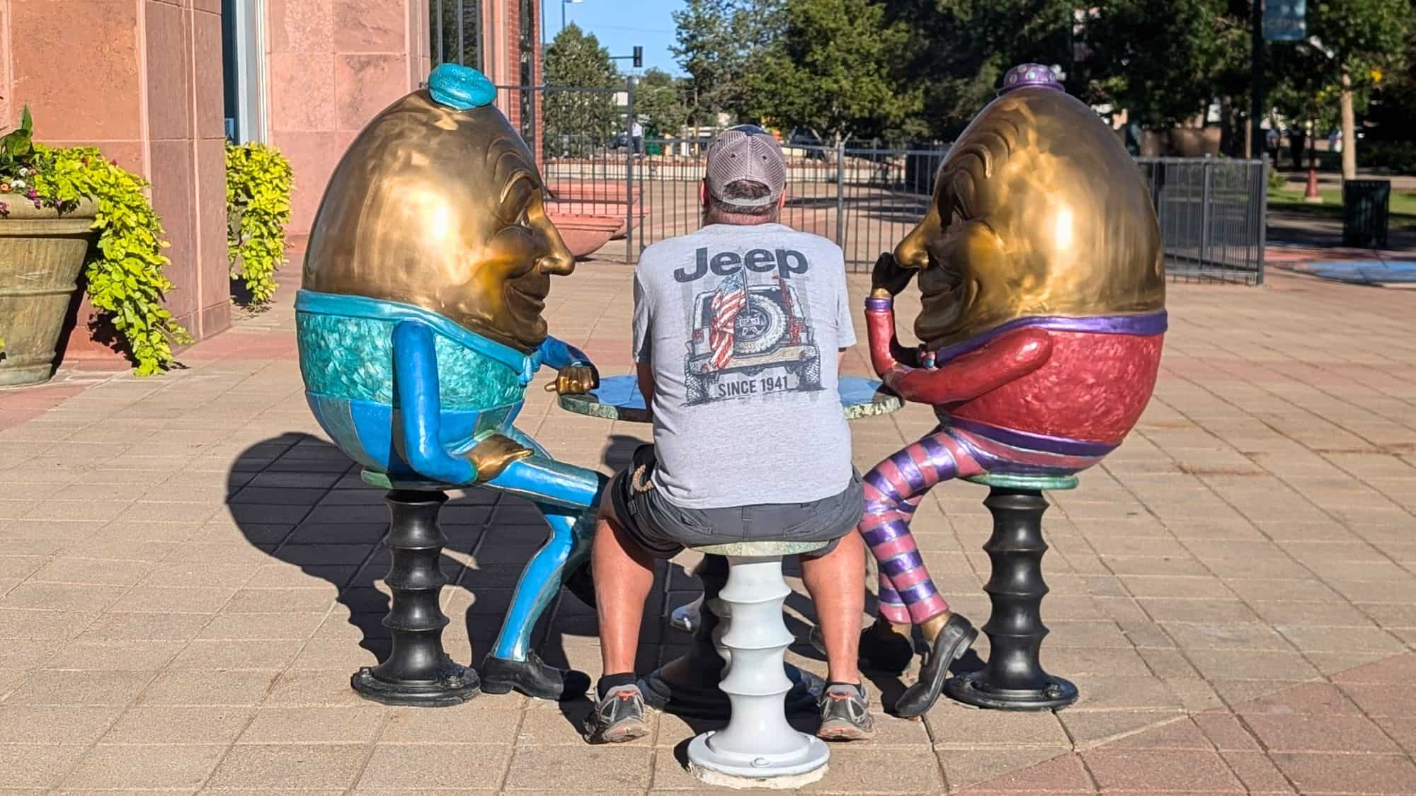 A whimsical outdoor sculpture of two large golden egg characters seated at a table, with a man in a Jeep t-shirt sitting between them in a playful street scene.