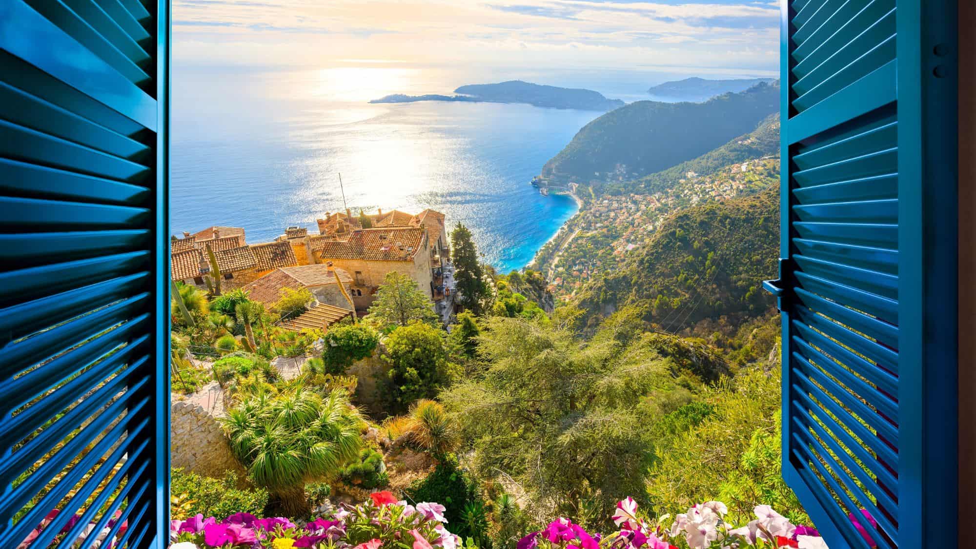 Eze, France - January 2 2024: View through an open window with blue shutters and flowers of the hilltop medieval old city of Eze, France, with the Mediterranean coastline and see below.