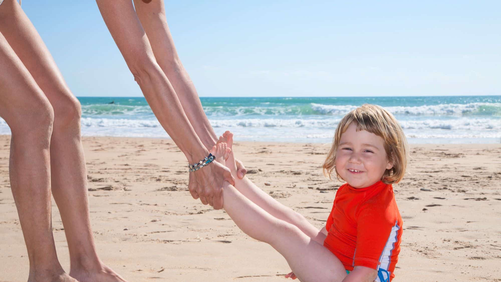 A child in an orange rash guard smiles while being swung by the feet on a sunny beach, with ocean waves and blue skies in the background.