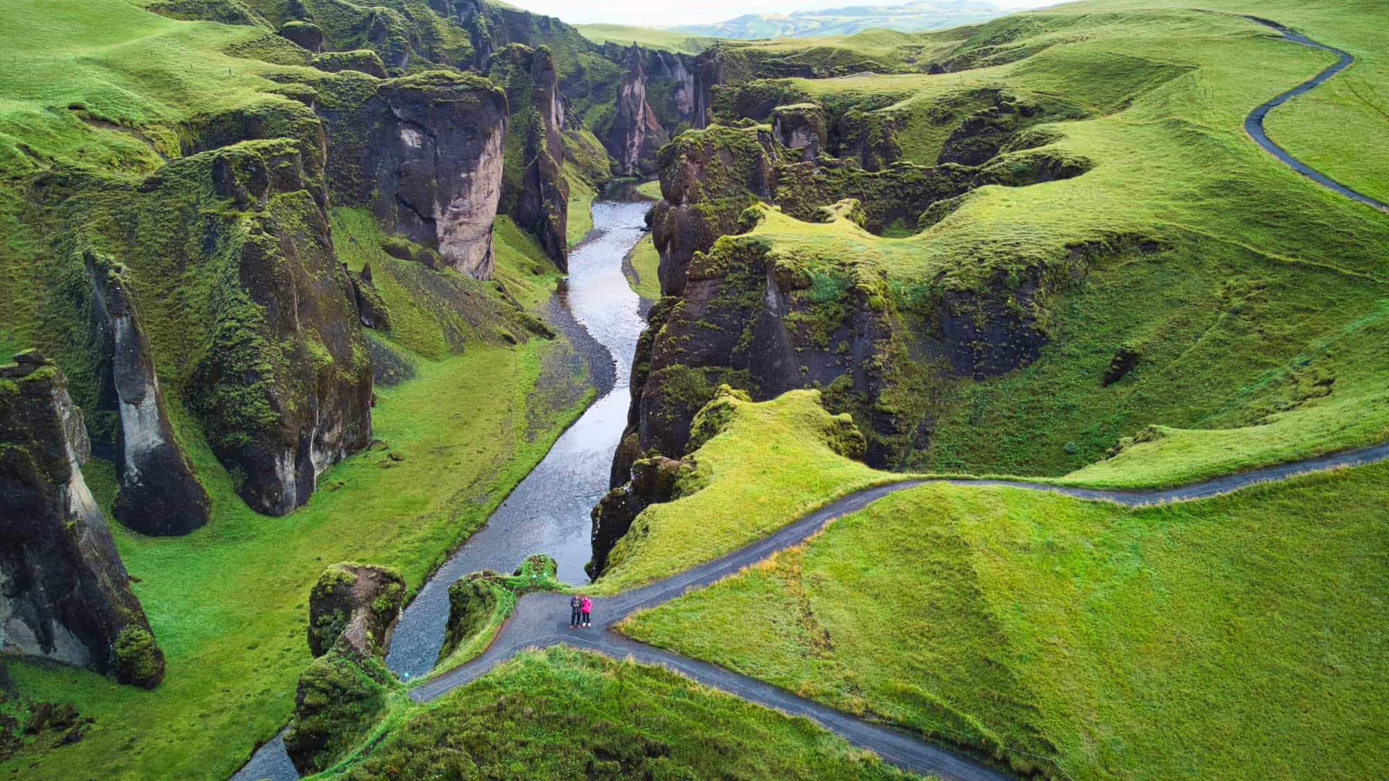 A winding river cuts through the dramatic moss-covered cliffs of Fjaðrárgljúfur Canyon in Iceland, with a couple standing on a path below, dwarfed by the towering green rock formations.