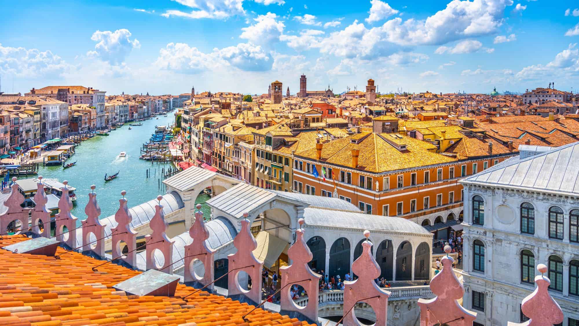 A stunning panoramic view over Venice from the Fondaco dei Tedeschi rooftop, showcasing the Grand Canal, the Rialto Bridge, and a sea of terracotta rooftops under a bright blue sky.