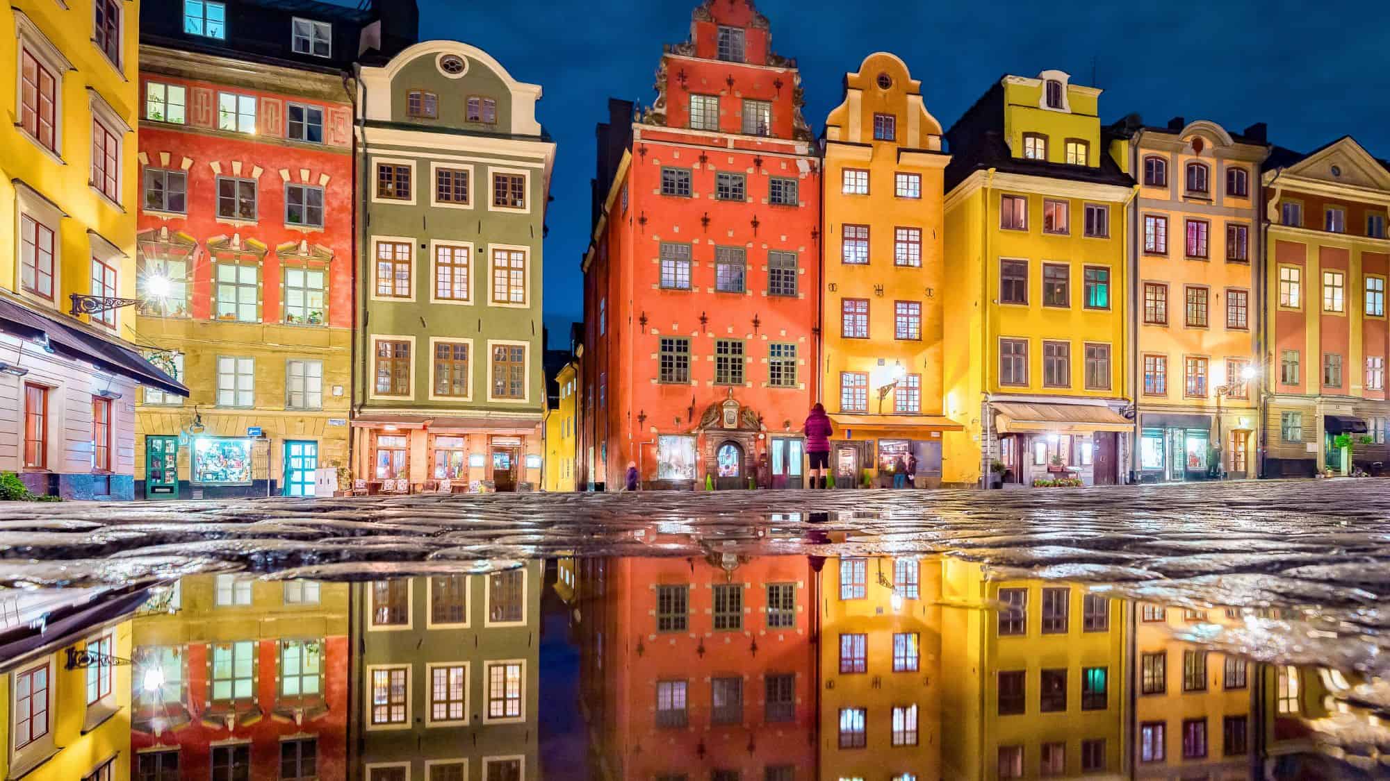 A row of colorful historic buildings in Stockholm’s Gamla Stan reflect in a puddle on the cobblestone square at night, glowing warmly under streetlights and a deep blue sky.