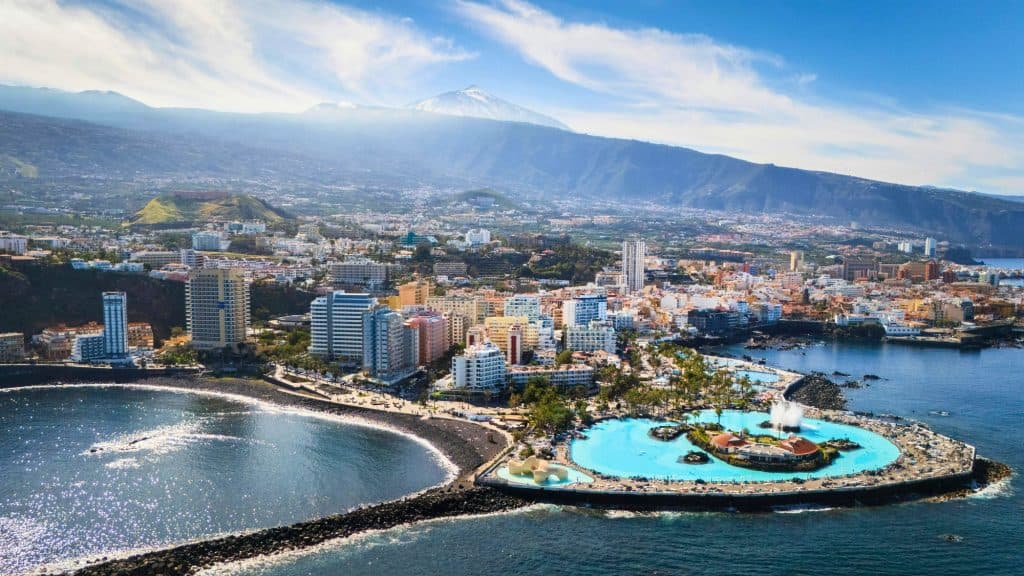 An elevated view of the colorful coastal town of Garachico in Tenerife, with red-roofed buildings, a white church tower, a sports complex, and rocky shoreline framed by the Atlantic Ocean and an offshore islet.