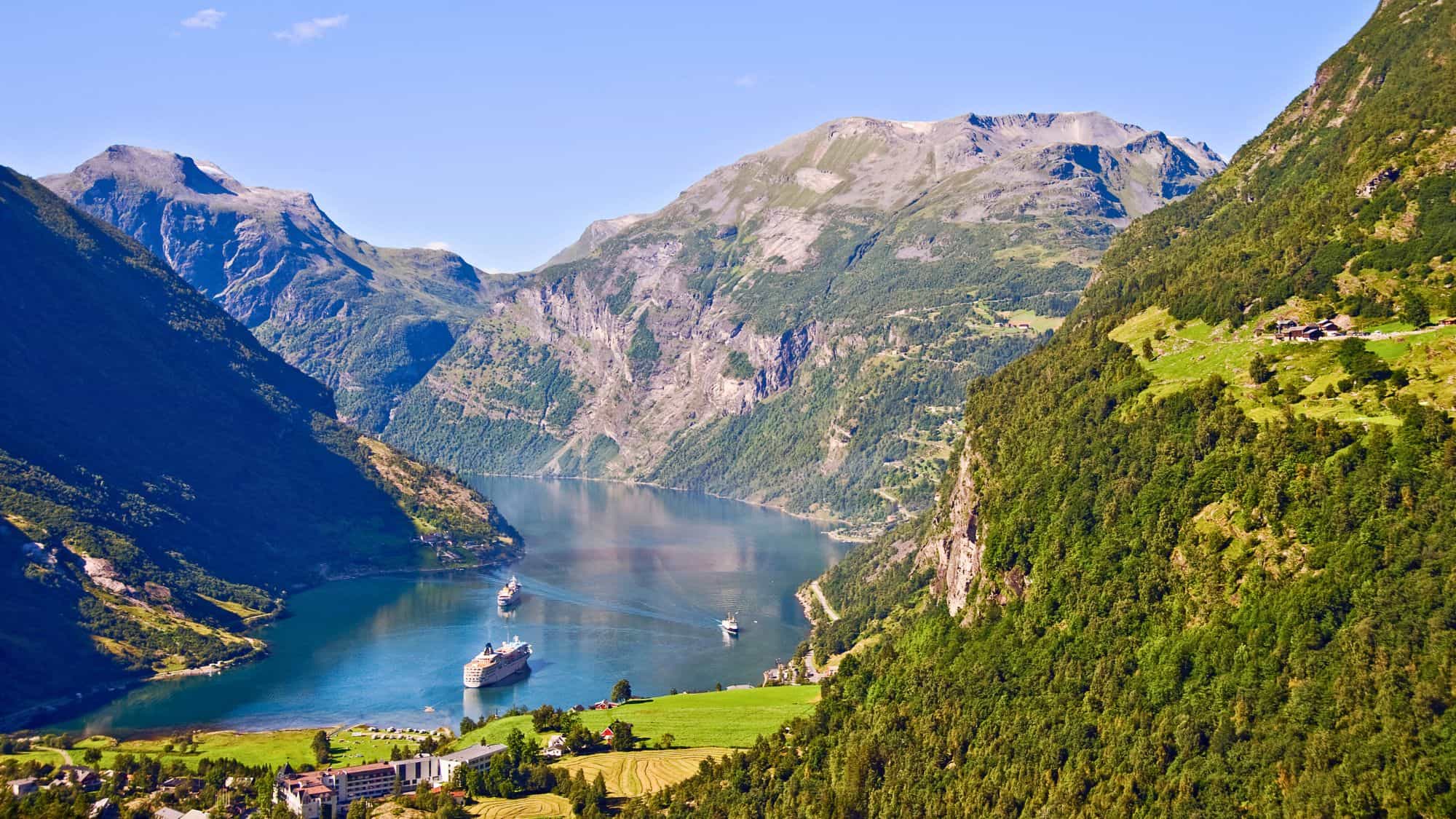 Cruise ships glide through the still, deep blue waters of Geirangerfjord, surrounded by steep, forested mountain slopes and scattered farmsteads in the Norwegian countryside.