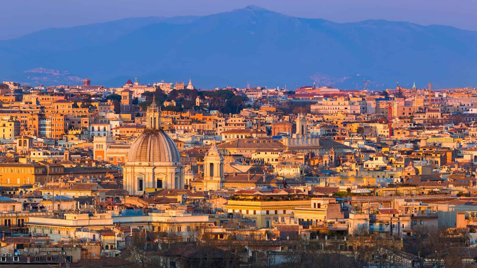 A panoramic shot of Rome from Janiculum Terrace at golden hour, with domes and terracotta rooftops bathed in warm light, backed by deep blue mountains under a lavender sky.