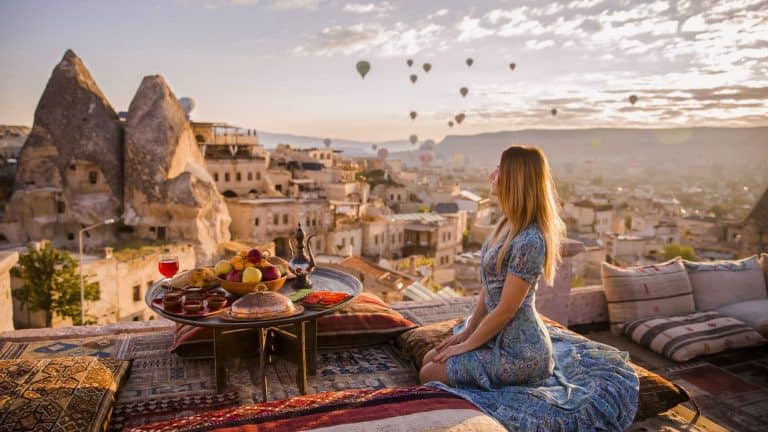 A woman in a blue patterned dress sits on a rooftop terrace in Göreme, Turkey, surrounded by colorful cushions and a table set with fruit, tea, and pastries, gazing at the iconic fairy chimneys and hot air balloons floating in the sunrise-lit sky.