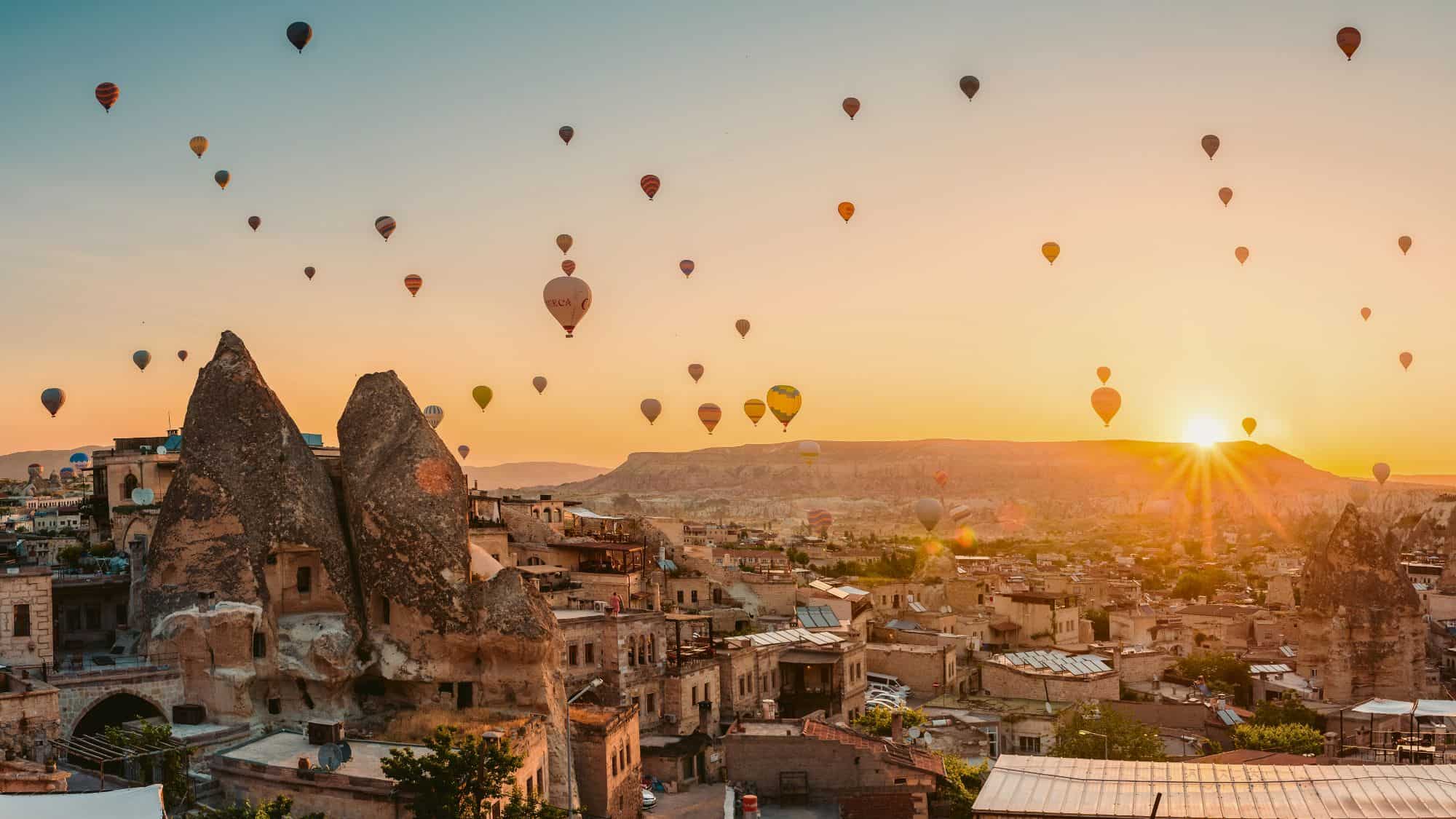 A surreal sunrise scene with dozens of hot air balloons floating over the unique rock formations and stone homes of Cappadocia.