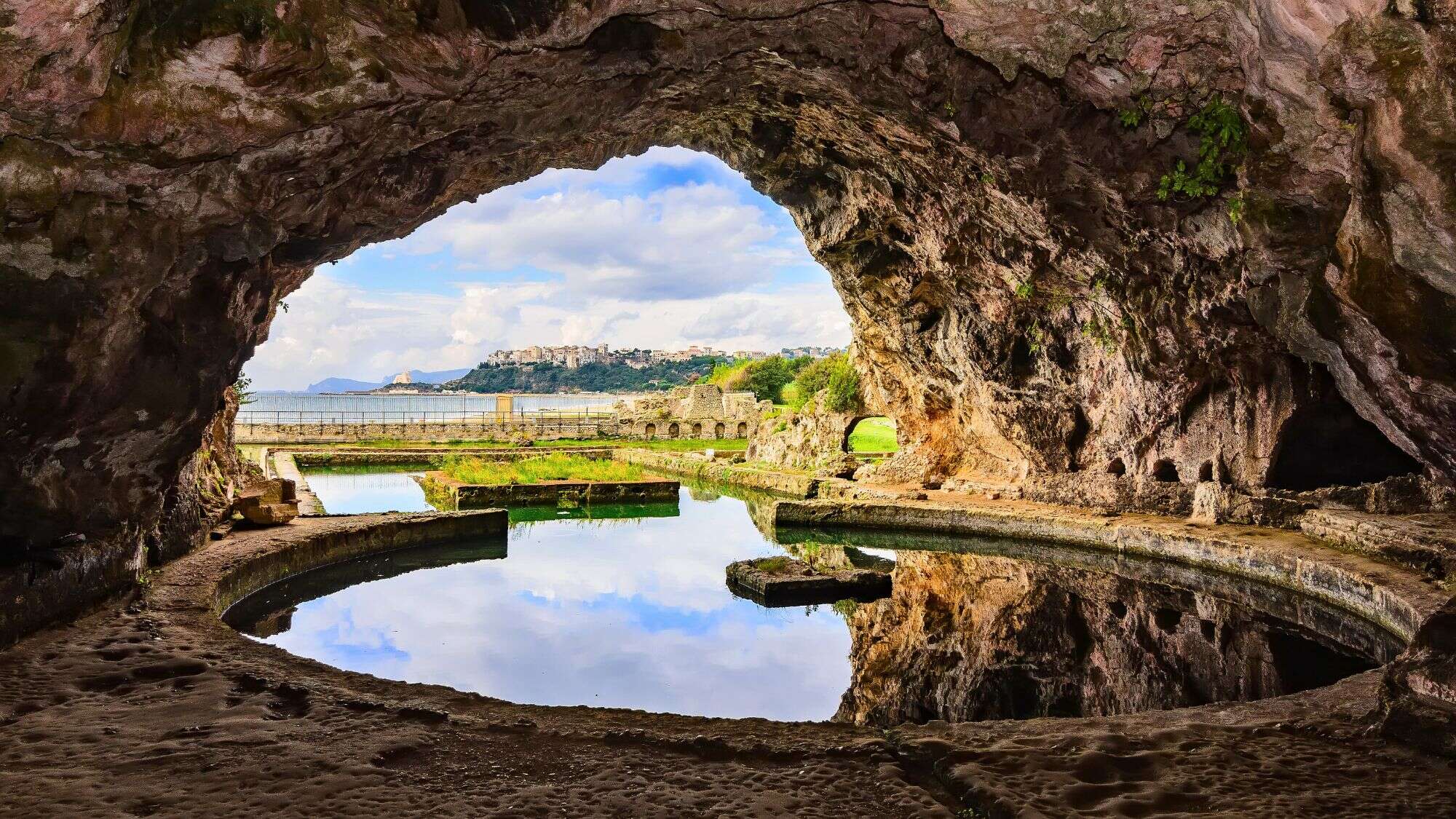 View from inside a coastal grotto looking out to a calm pool and ruins with scenic coastal cliffs and a distant village framed by the cave opening.