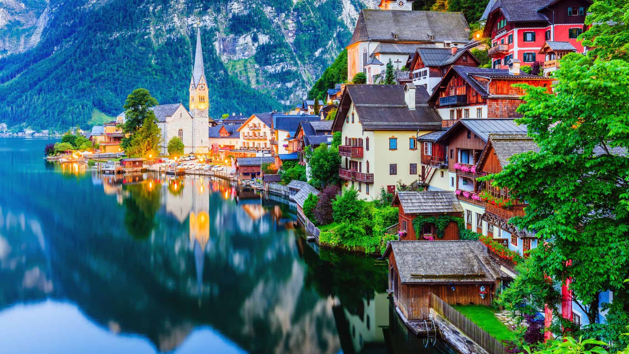 A picturesque lakeside village with pastel buildings and a tall church spire, nestled between serene waters and dramatic alpine mountains.