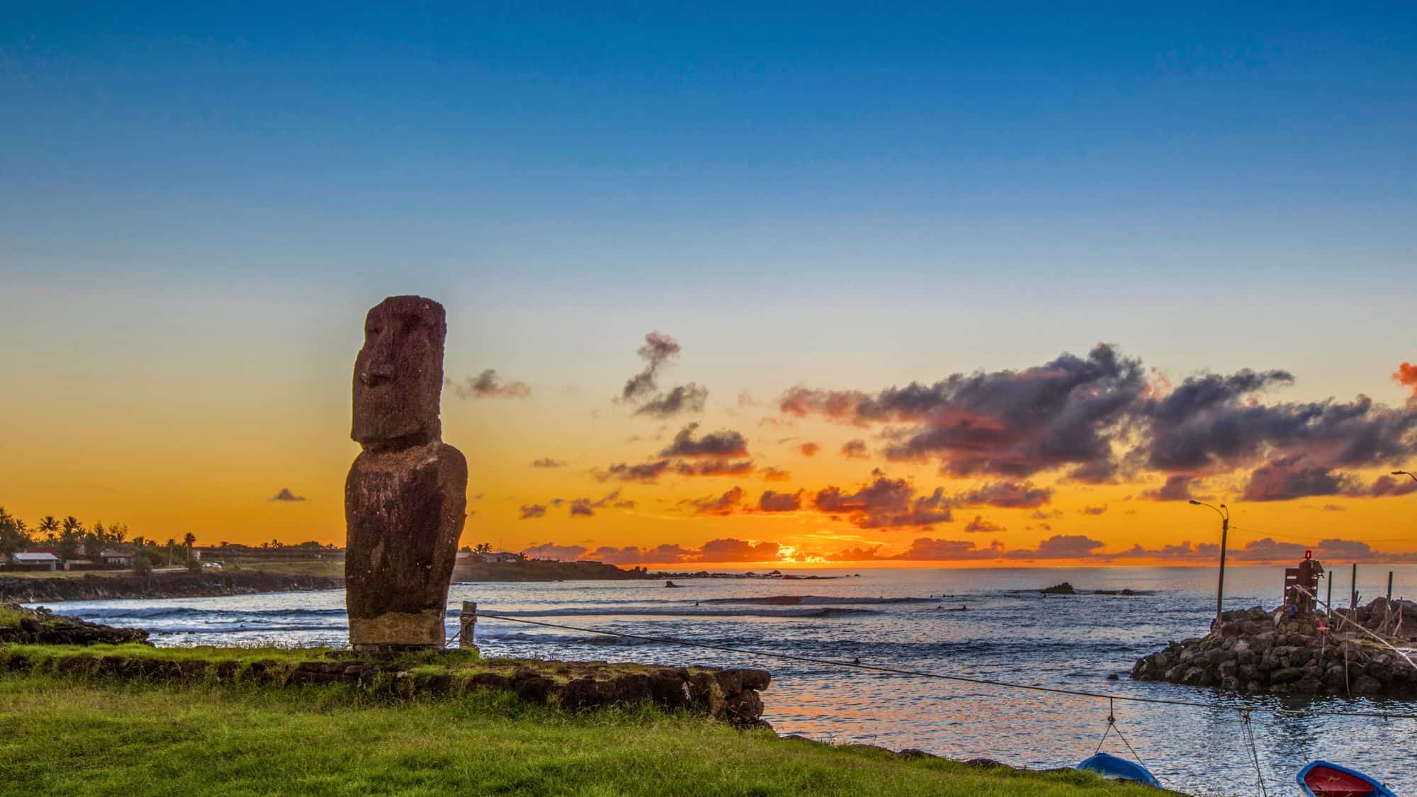 A solitary Moai statue stands tall near the coastline of Easter Island as the sun sets, casting an orange and pink glow across the sky and reflecting on the calm ocean. The scene blends ancient culture with natural beauty.