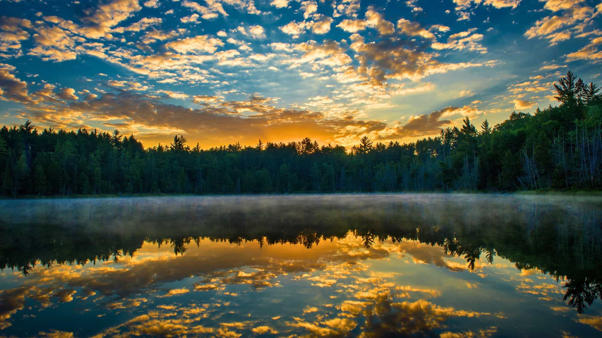 Golden sunrise peeks over a dense pine forest at Hartwick Pines State Park, casting a fiery glow across scattered clouds and mirroring perfectly on the still, misty lake below.