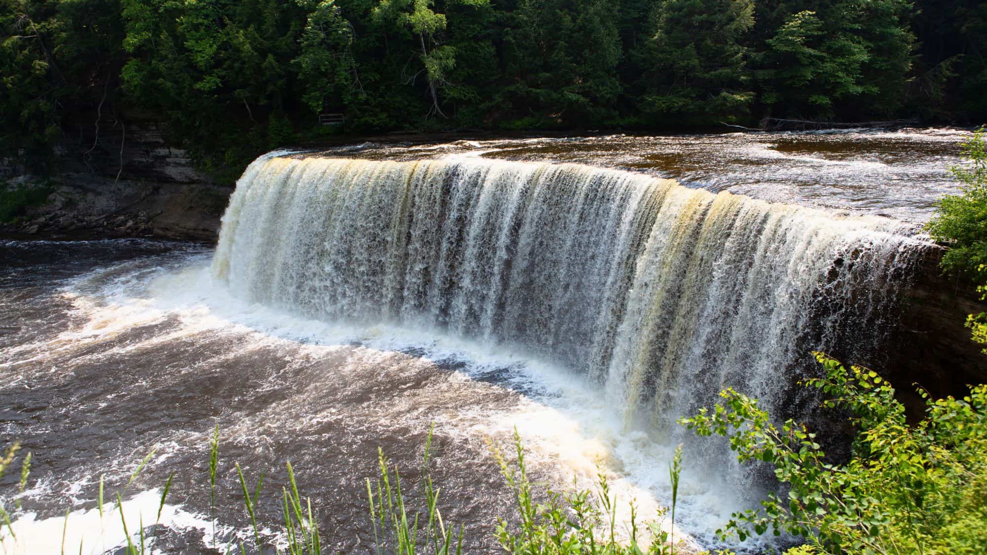 Wide, powerful waterfall cascades over a rock ledge surrounded by lush forest, with frothy water plunging into the brown river below.