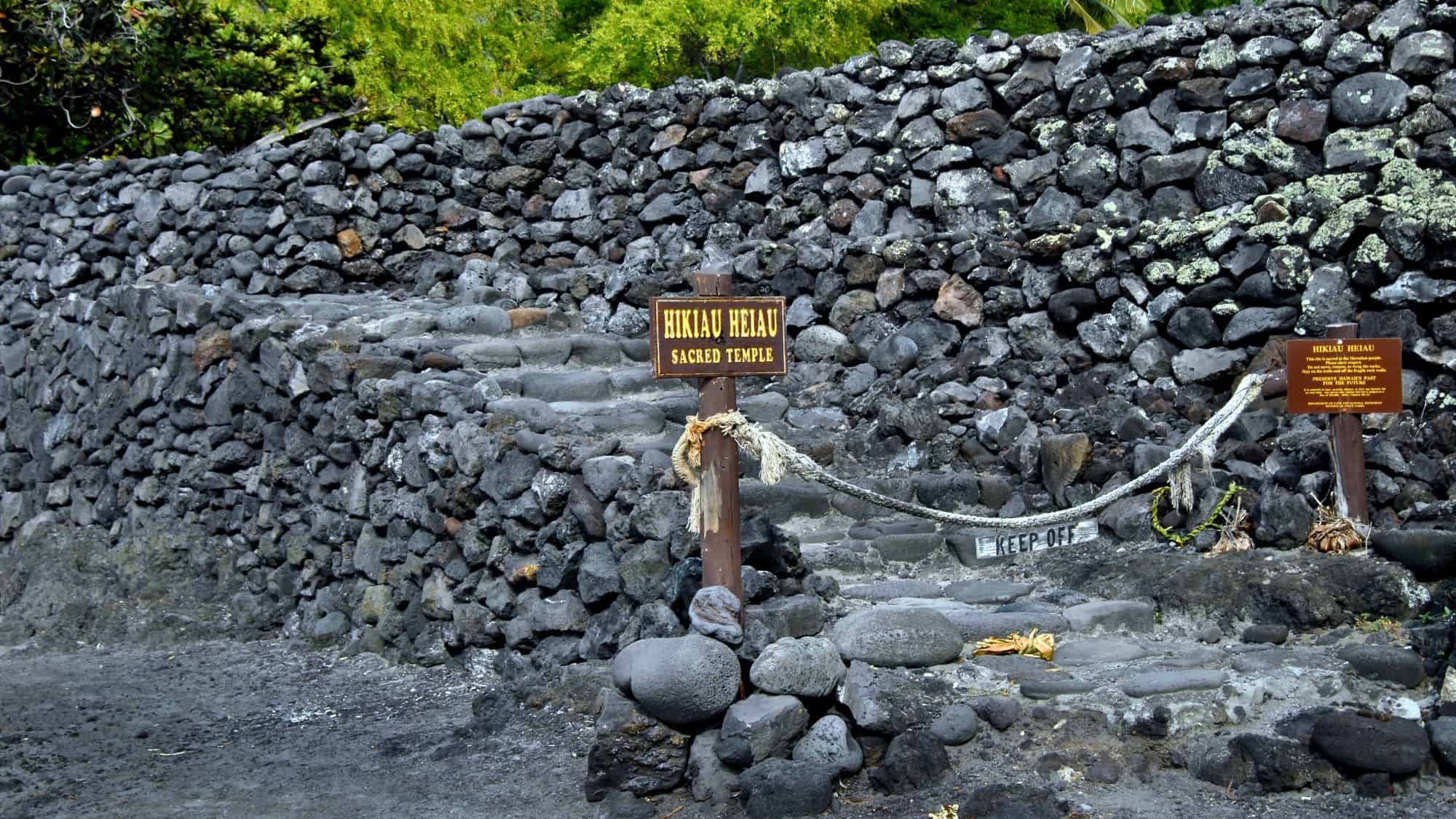 Stone ruins of the Hikiau Heiau sacred temple in Hawaii, with signs designating its cultural significance and rope barriers requesting visitors to keep off.