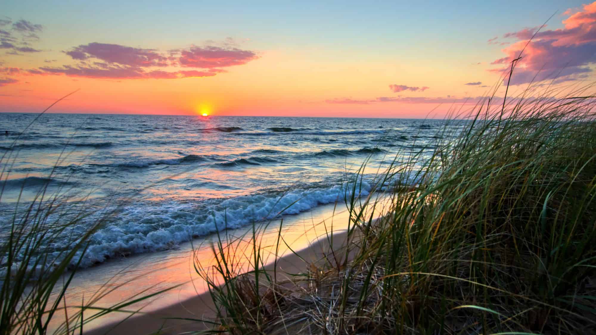 Vibrant sunset over Lake Michigan, with pink and orange skies reflecting on gentle waves and tall grasses lining the sandy shore.