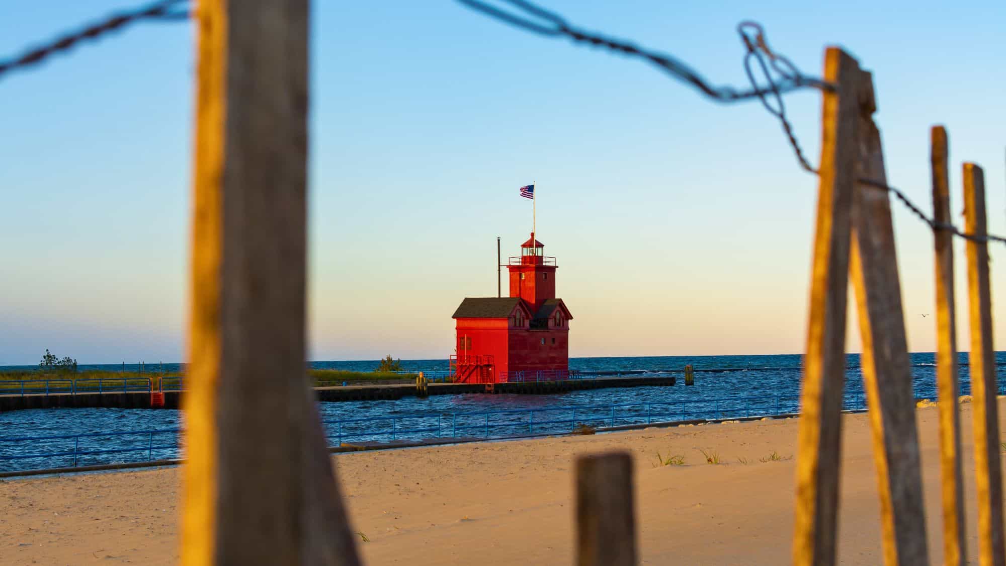 Iconic red lighthouse known as Big Red stands at the end of a pier, framed by a sand fence and backlit by the warm glow of evening light.