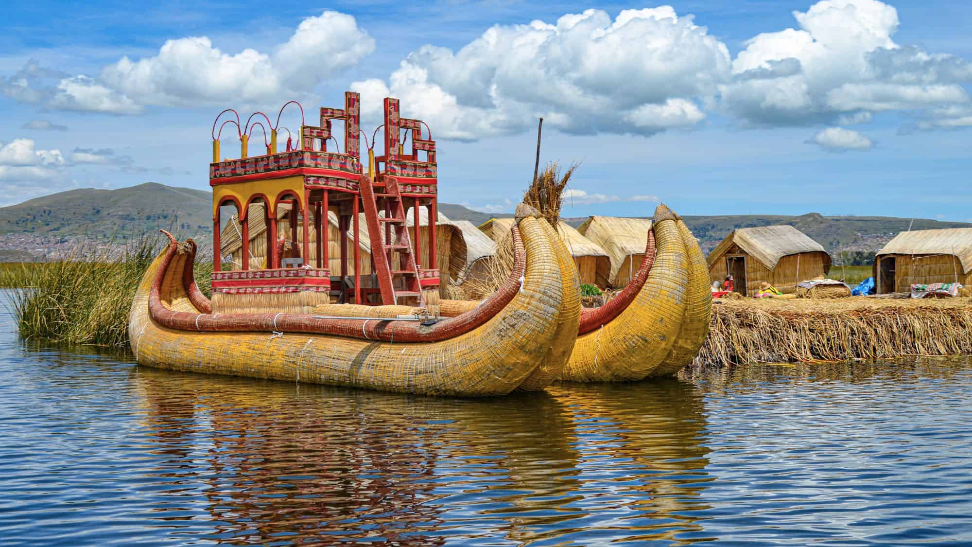 A traditional reed boat with a red wooden deck floats near the edge of Lake Titicaca, surrounded by thatched huts and tall grasses. The scene showcases Indigenous craftsmanship and life on the Uros floating islands.