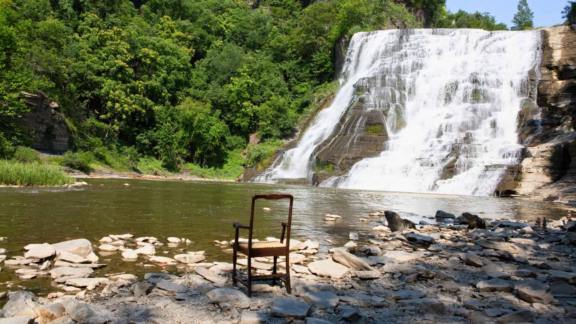 A powerful waterfall fans out over a steep, rocky ledge into a calm pool below, with a single wooden chair oddly placed on the rocky shoreline in the foreground.