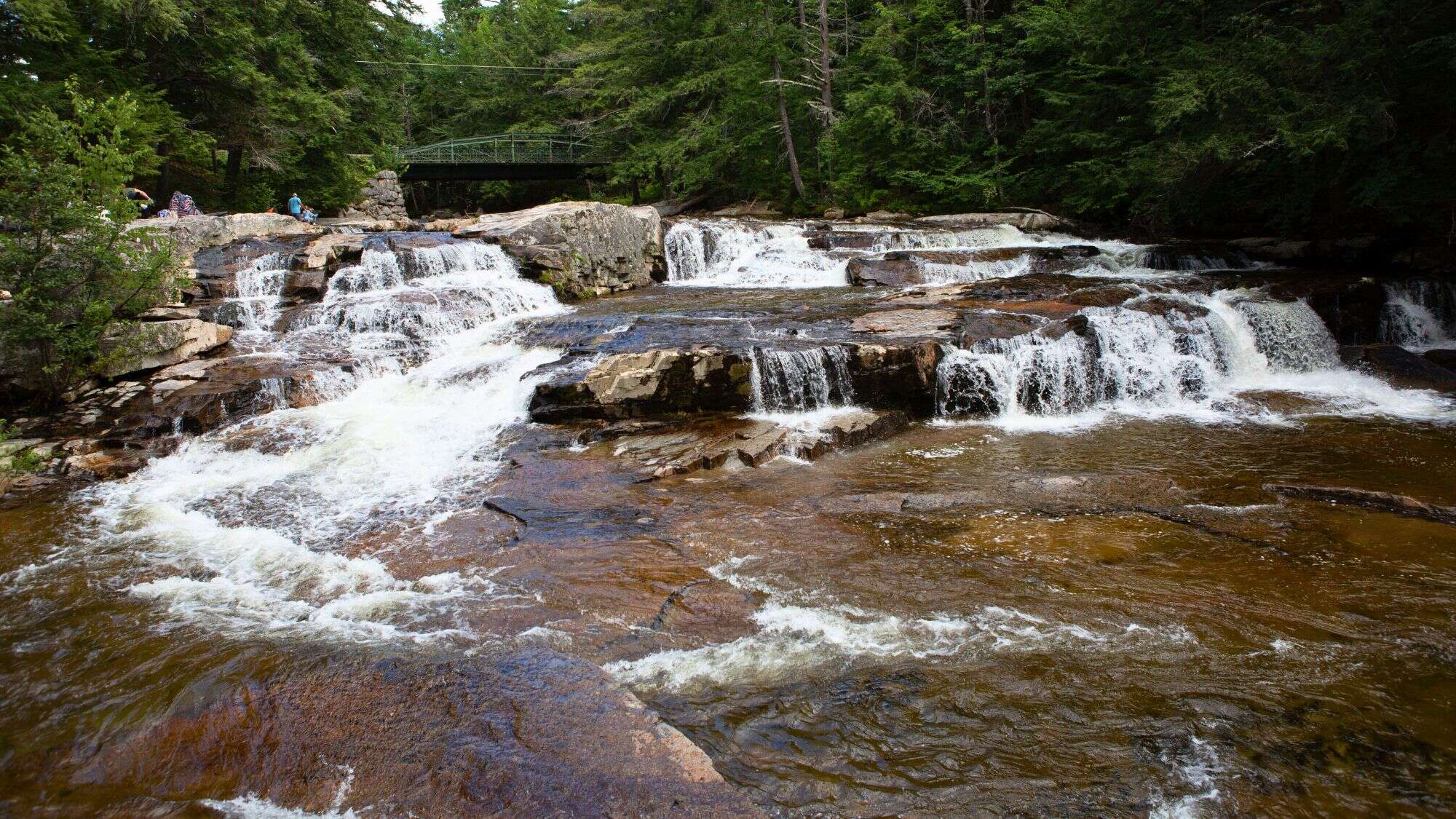 A scenic cascade tumbles over layered rock ledges beneath a green metal bridge, with people lounging and wading near the falls amid dense trees.