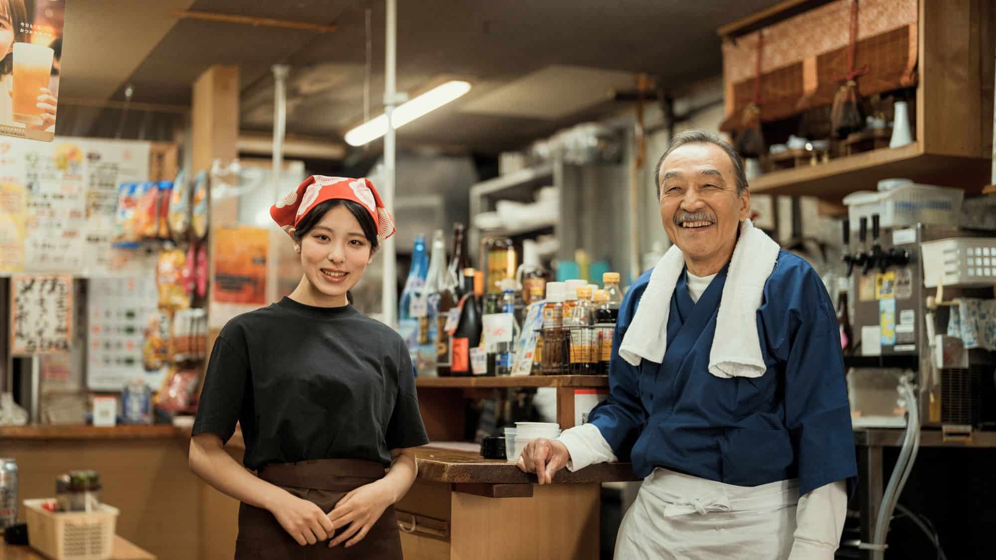 A smiling man and woman stand in a cozy Japanese restaurant, dressed in traditional service attire with a welcoming ambiance full of sauces and signage in the background.