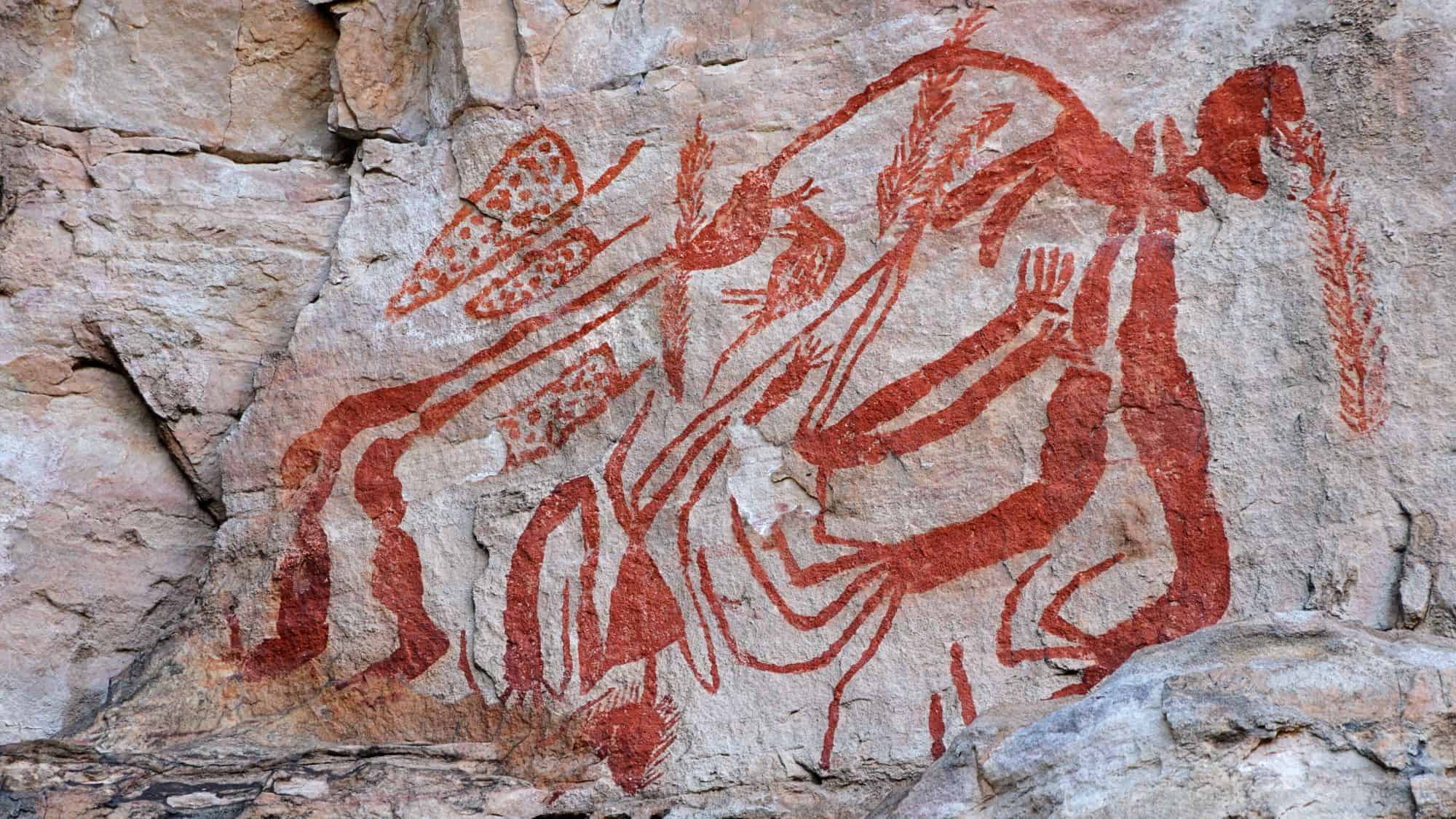 Aboriginal rock art painted in vibrant red hues decorates a sandstone cliff, showing human figures and animals in intricate patterns. The ancient artwork stands as a cultural testament to Australia’s Indigenous heritage.