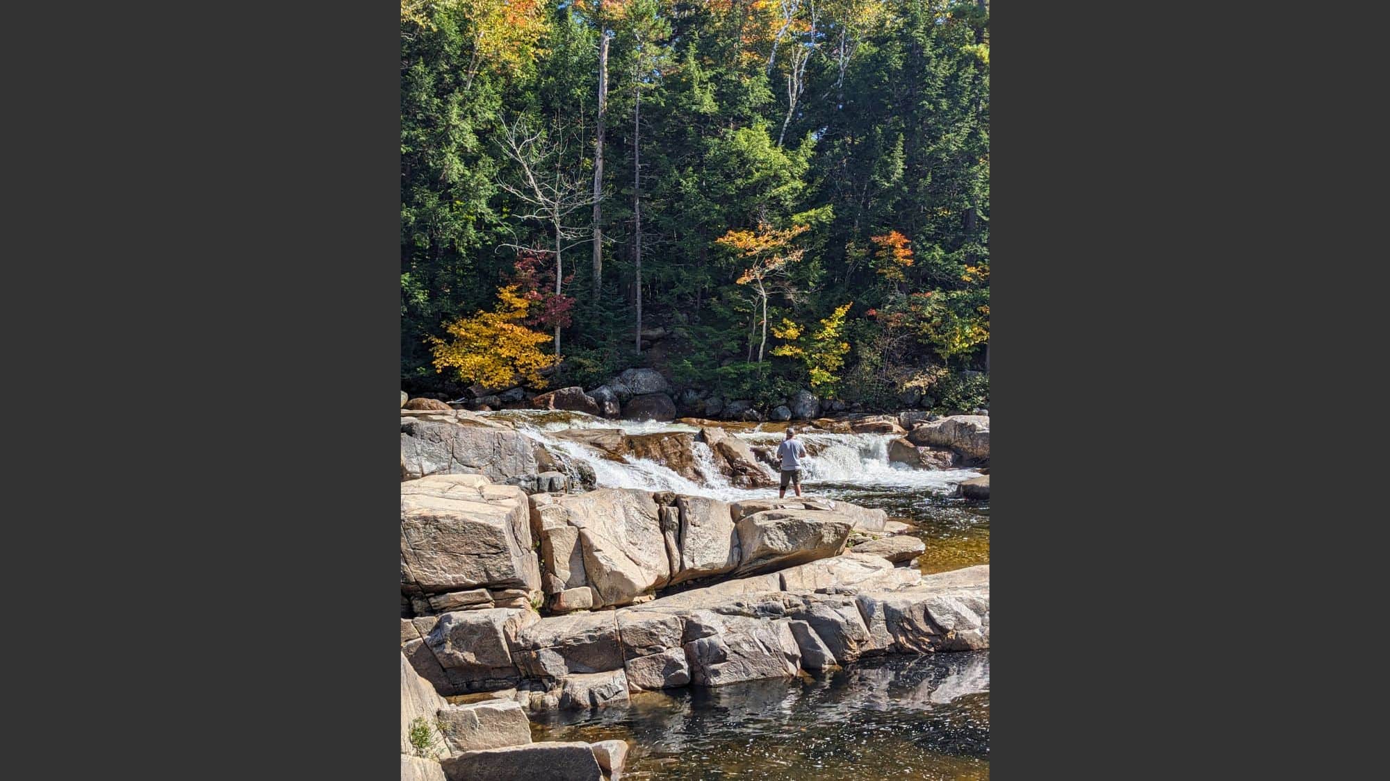 A person stands fishing on large boulders at the base of a small waterfall, surrounded by vivid autumn trees and a serene, forested backdrop.