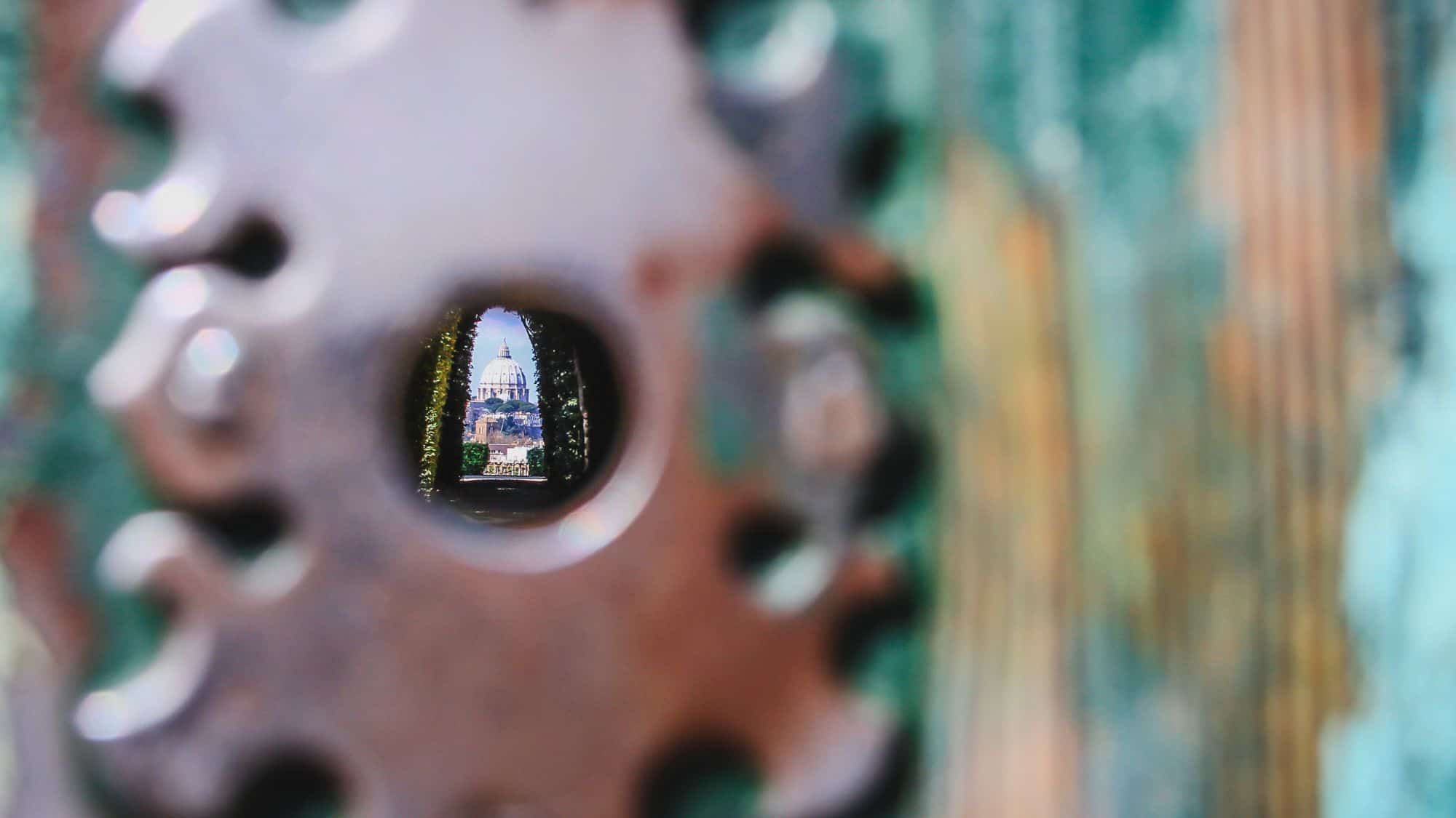 A perfectly aligned view of St. Peter’s Basilica in the Vatican appears framed through the famous keyhole on the Aventine Hill in Rome, surrounded by greenery and the metallic edge of the keyhole.
