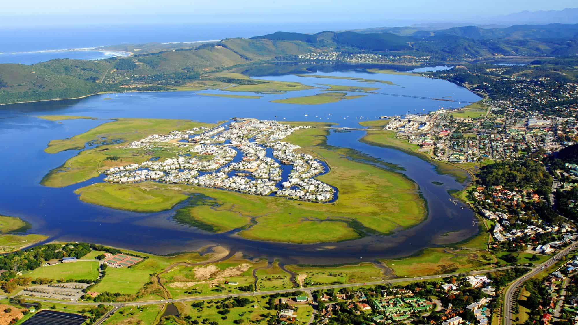An aerial shot captures the lush lagoon and estuary system surrounding the town of Knysna, South Africa. Winding waterways snake through green marshlands and modern developments, with the Indian Ocean in the distance.