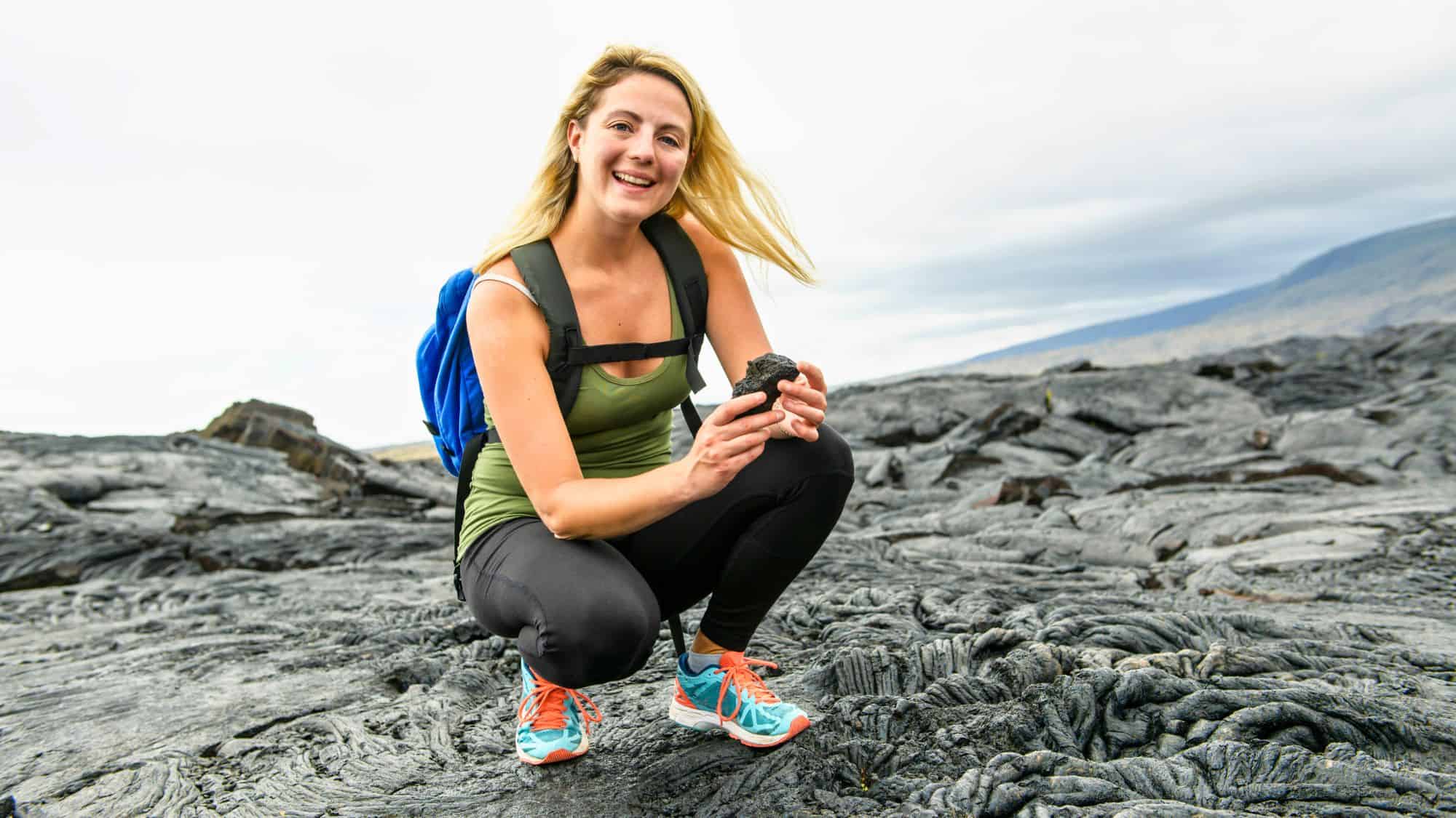 A smiling hiker crouches on black lava rock terrain, holding a chunk of volcanic rock, with rugged formations and distant hills under an overcast sky.