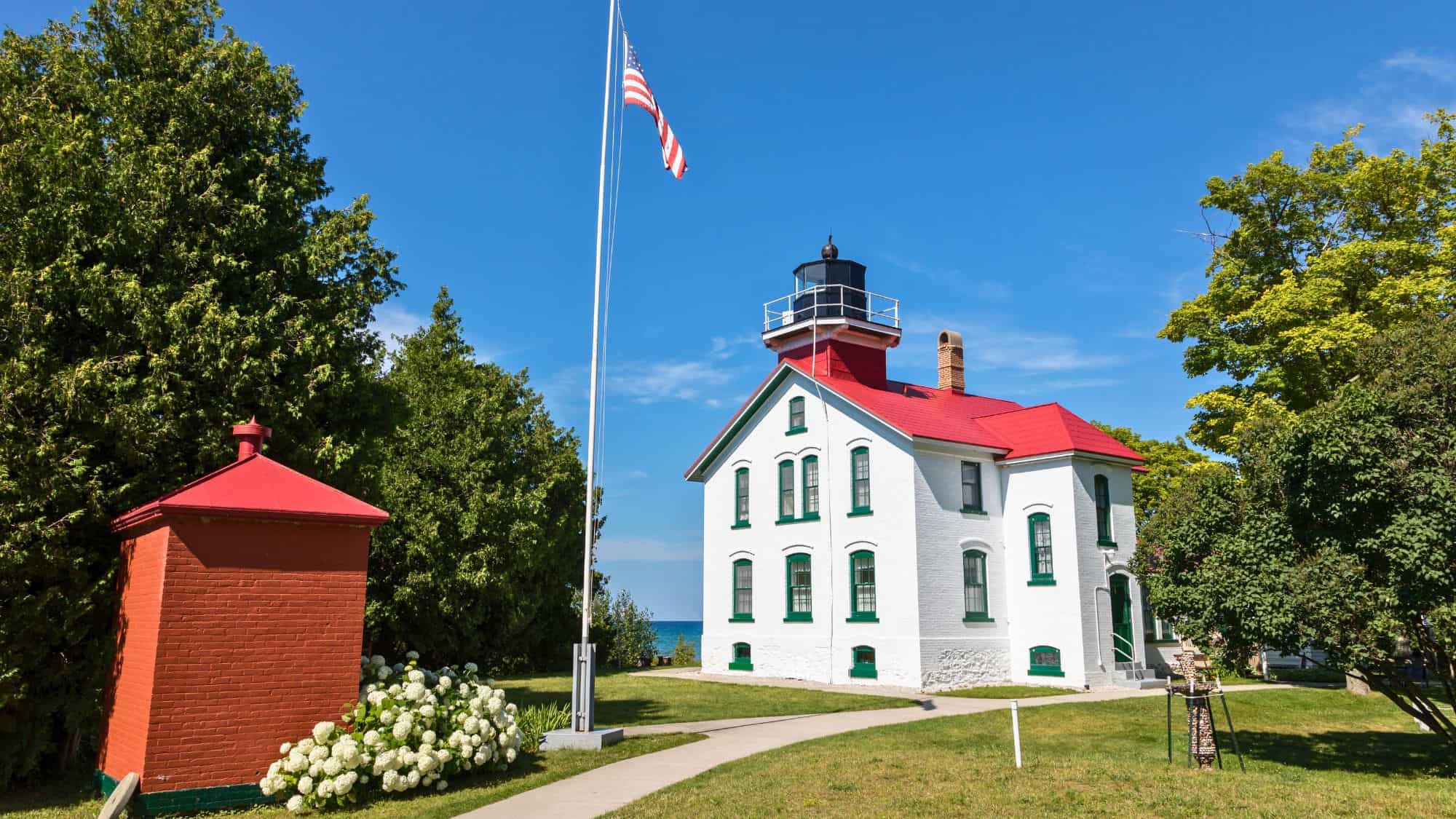 Historic white lighthouse with red roof and green trim, surrounded by manicured lawn, an American flag, and dense green trees under a bright blue sky.