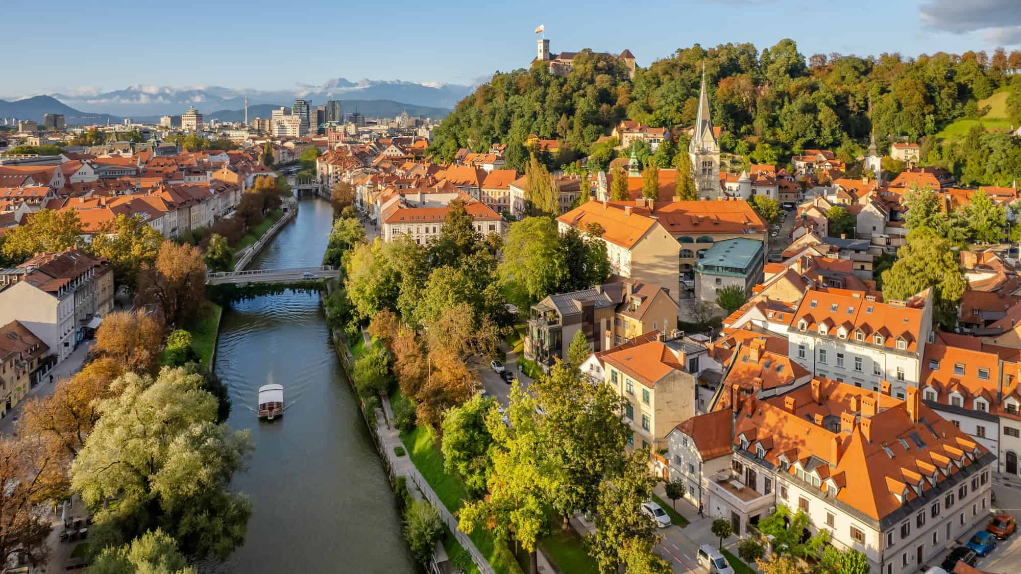 A scenic aerial of Ljubljana with the Ljubljanica River winding through the city, lined with leafy trees and orange-roofed buildings, while Ljubljana Castle sits perched on a hilltop surrounded by forested slopes.