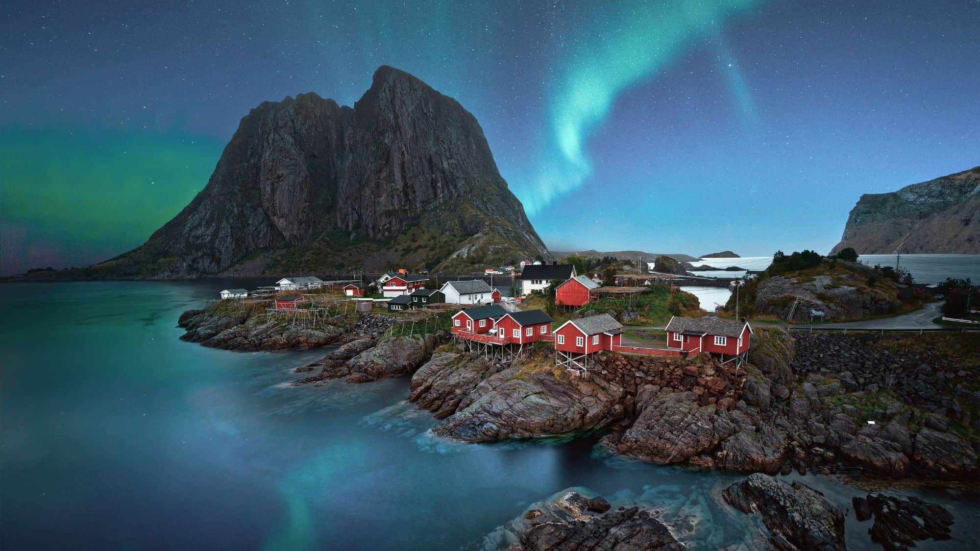 Red fisherman cabins sit on rocky shores under a jagged mountain peak, with the Northern Lights glowing green above the peaceful village in the Lofoten Islands.