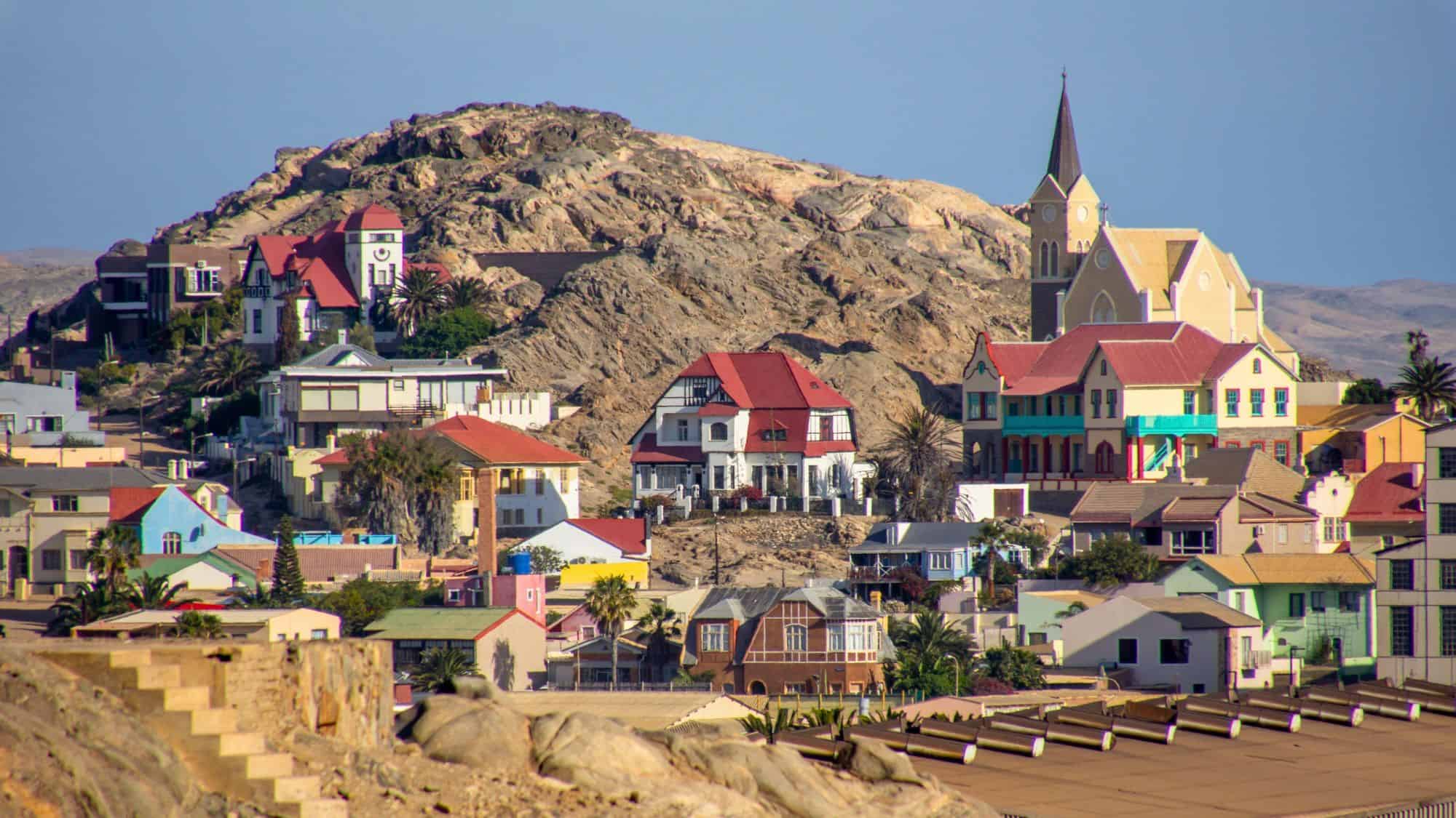 Colorful colonial-style buildings with red roofs and a prominent church spire sit clustered on a rocky hill in the coastal town of Lüderitz, Namibia. The arid backdrop contrasts with the lively architecture, evoking a mix of German heritage and desert landscape.